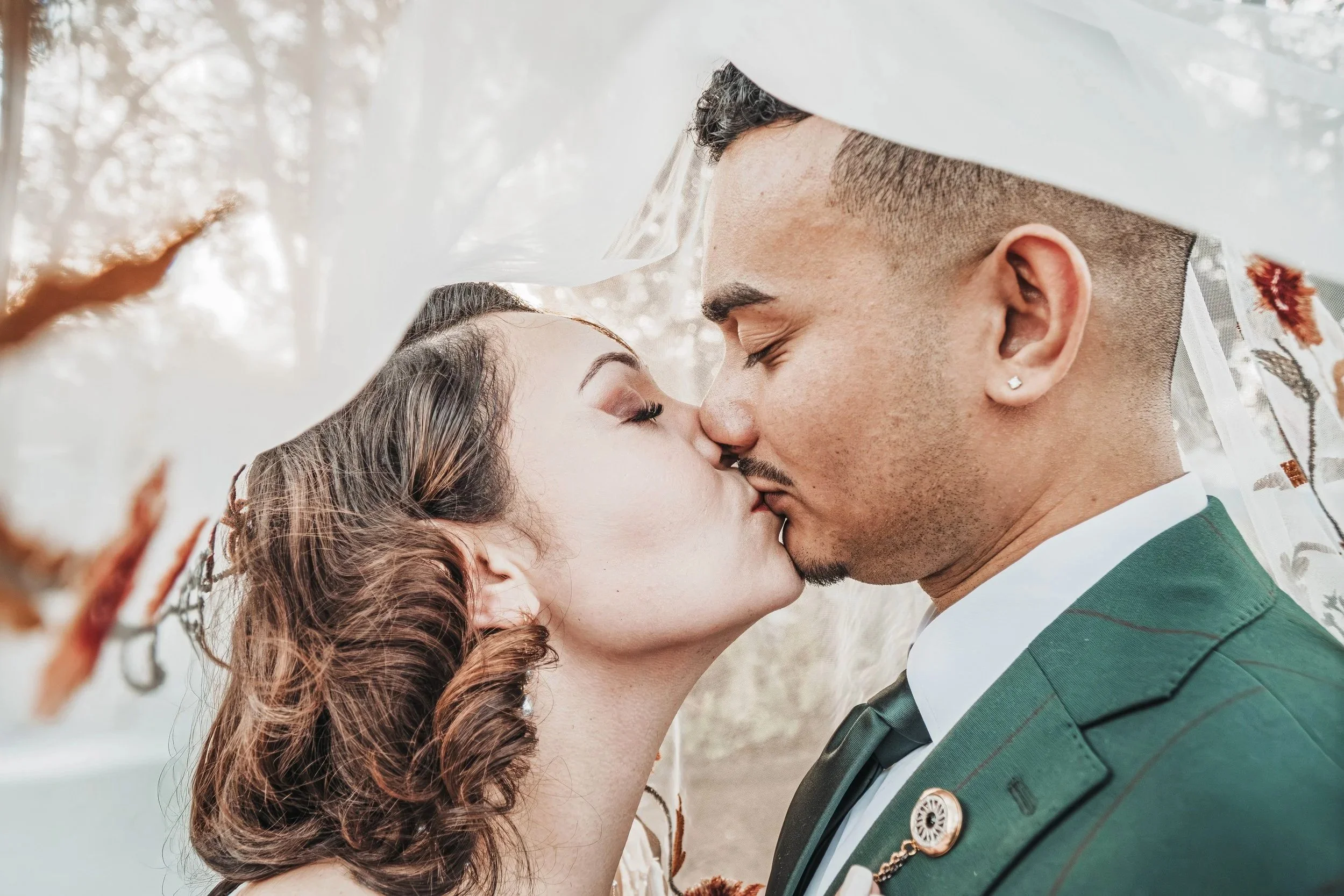 A close-up of an engaged couple about to kiss, with a white veil partially covering their faces, outdoors with blurred trees in the background.