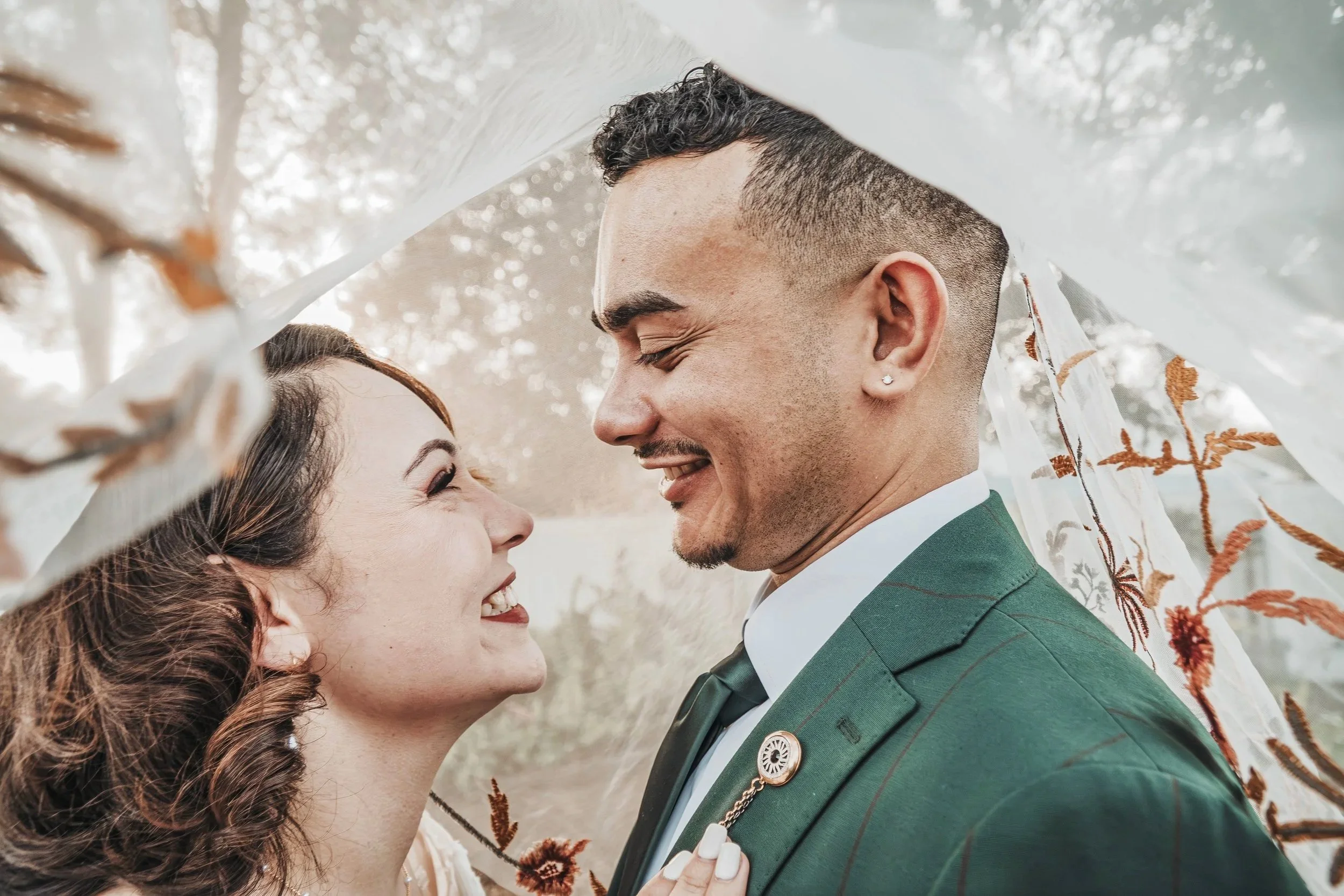 A happy couple outdoors, smiling, close together under a fabric canopy with embroidered orange and white designs.