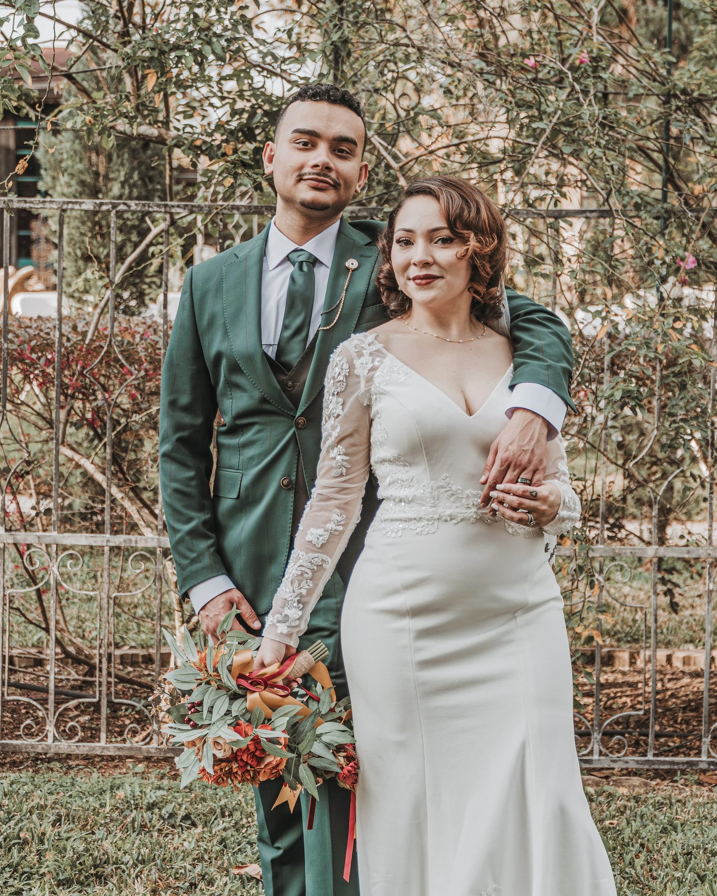A couple dressed in wedding attire standing outdoors in front of a metal fence with greenery and trees in the background. The man is wearing a green suit with a matching tie and a brooch, holding a bouquet of flowers. The woman is wearing a white wed