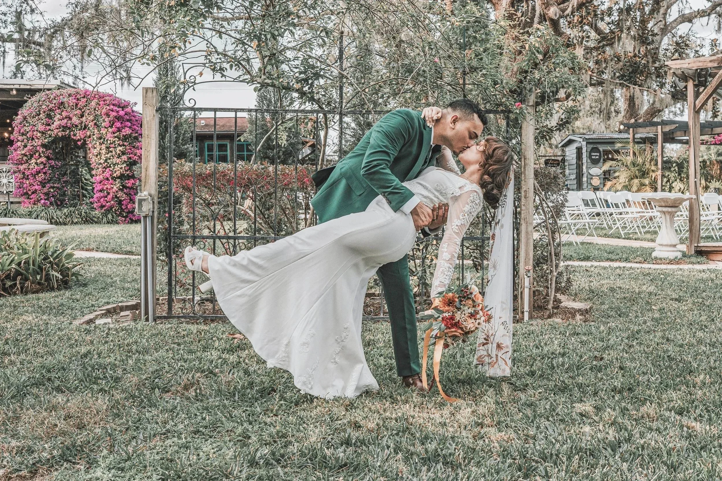 A man in a green suit dips a woman in a white wedding dress for a kiss in a garden setting with trees, purple flowers, and outdoor seating in the background.