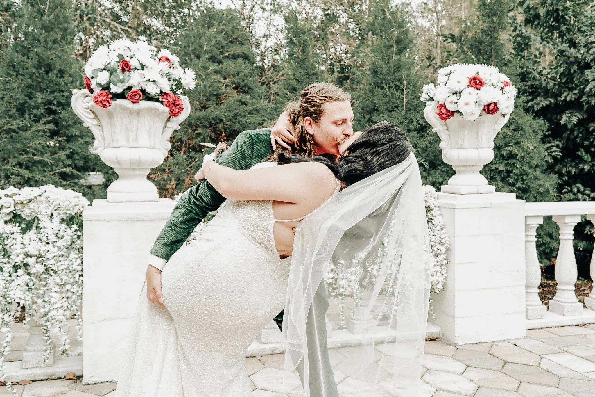 A person in a wedding dress hugging a person in a green jacket and kissing them on the lips, with a backdrop of large floral arrangements and green trees.
