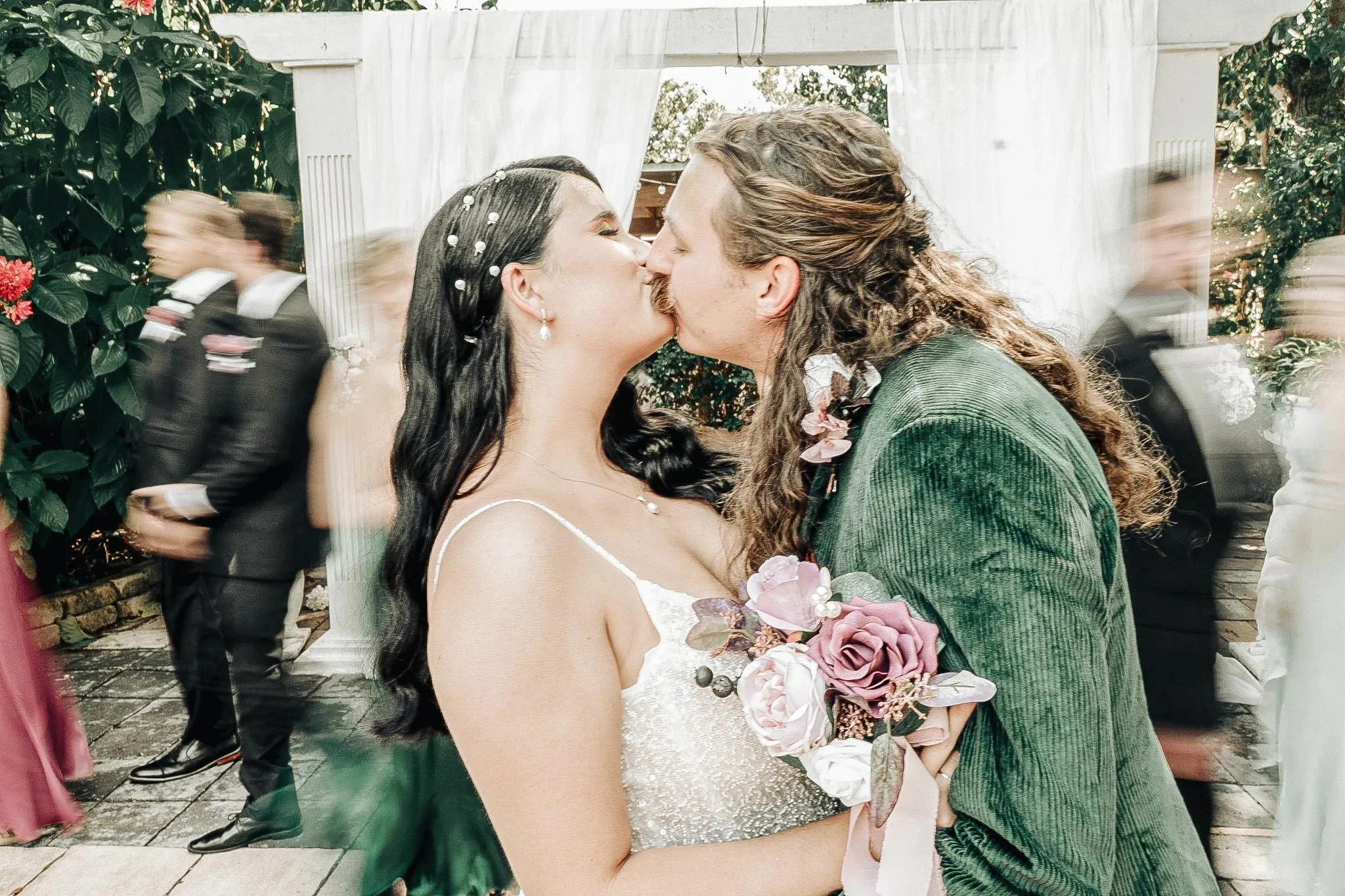 A bride and groom share a kiss at their wedding reception, surrounded by guests. The bride wears a white dress and pearl accessories, and the groom wears a green suit with floral boutonniere. The scene is outdoors with greenery and white drapery.