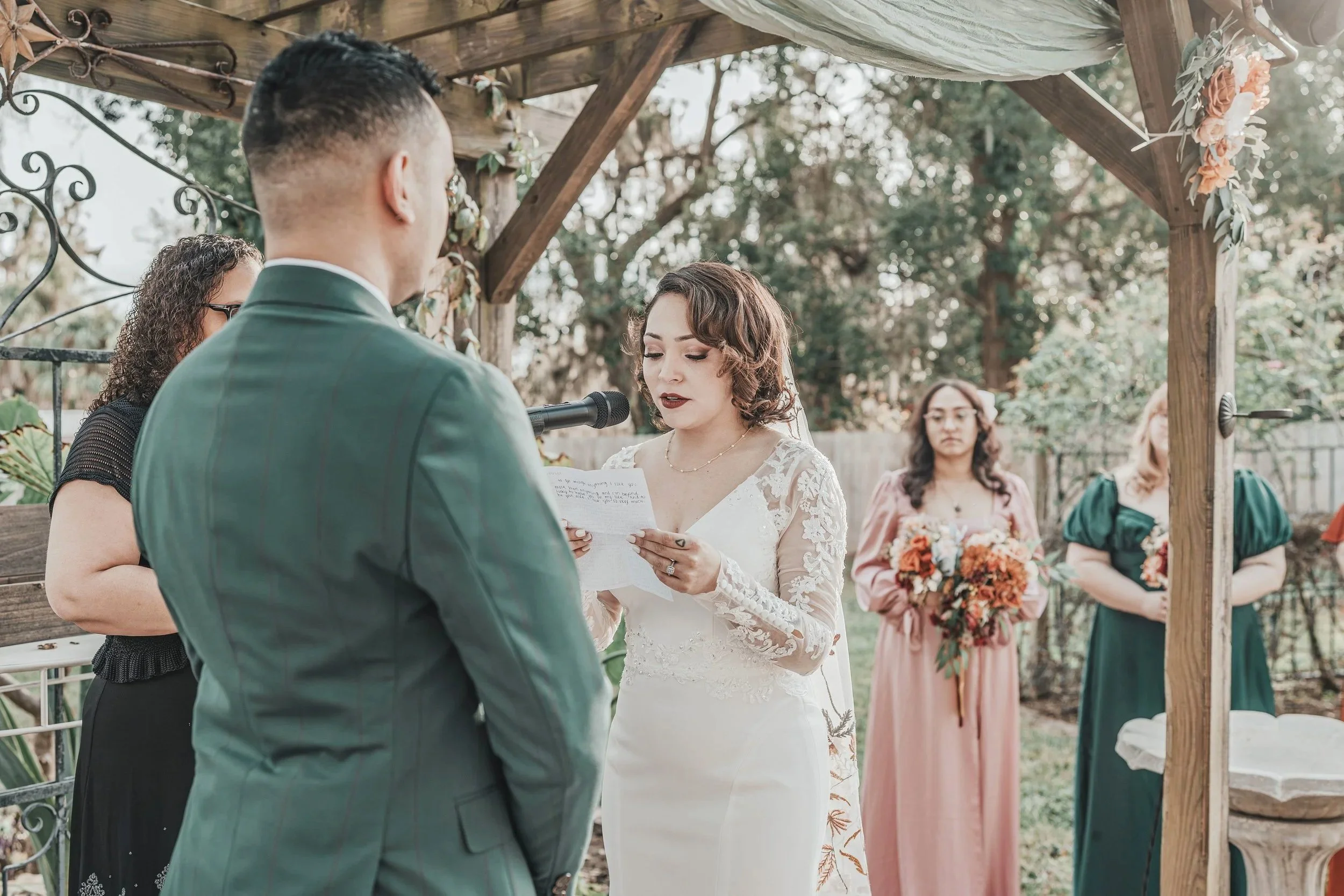 A woman in a white wedding dress reads vows during an outdoor wedding ceremony under a wooden arch decorated with flowers, with bridesmaids standing behind her holding bouquets.