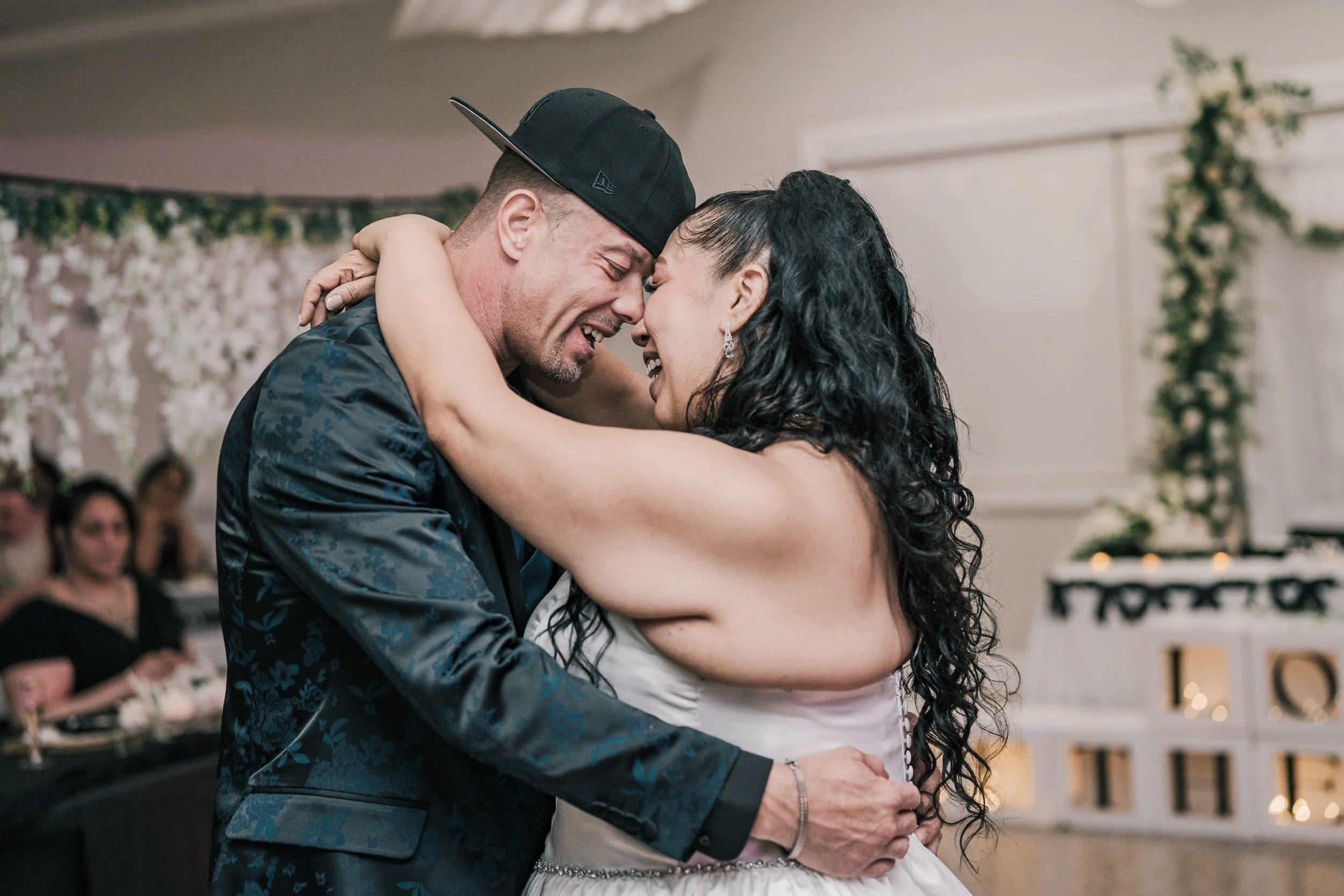 A couple, a man and a woman, share an intimate dance at a wedding reception, smiling with their foreheads touching. The man is wearing a black cap backward and a patterned shirt, and the woman has long dark curly hair and jewelry. In the background, 
