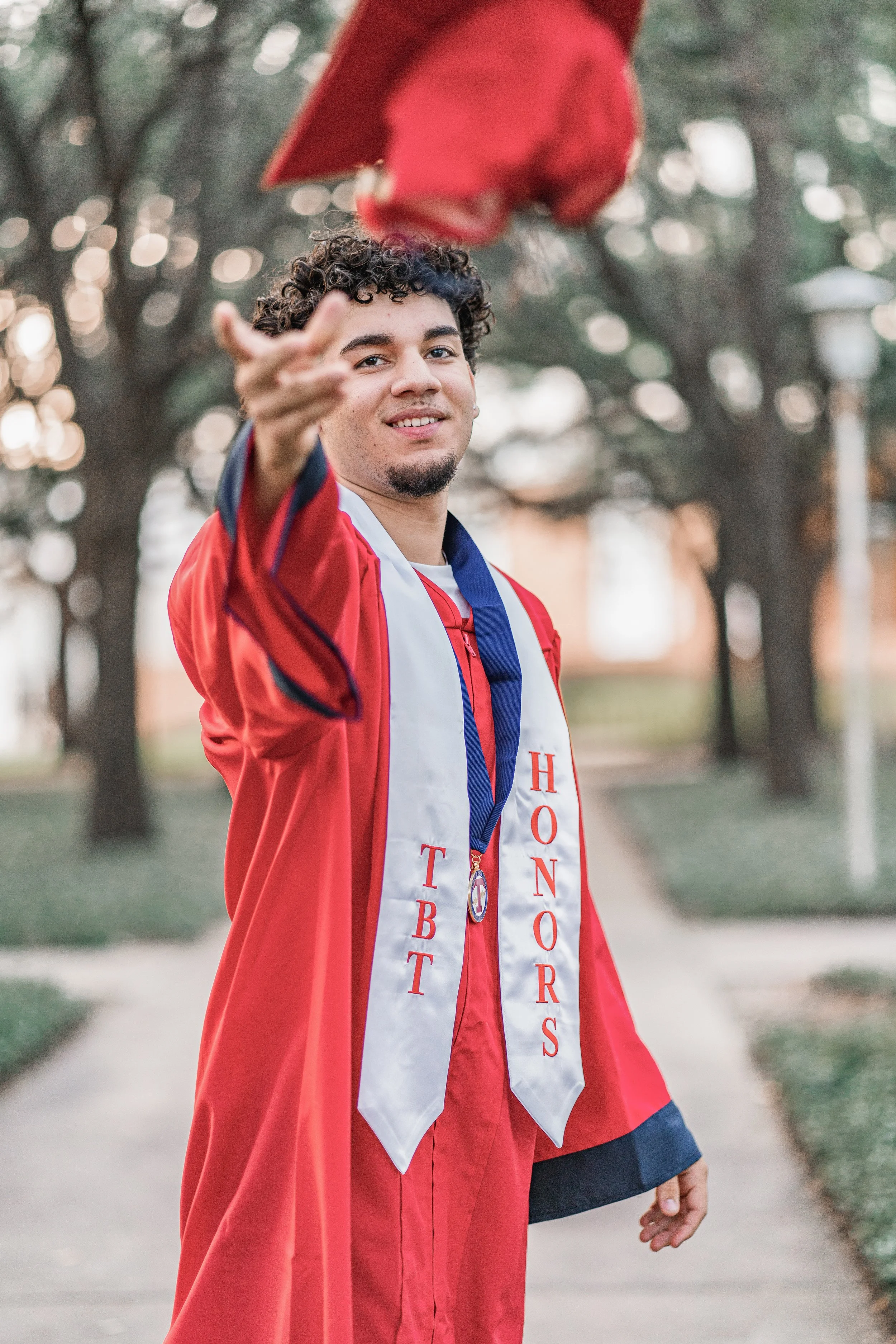A young man in a red graduation gown and white stole with red lettering, standing outdoors, smiling and pointing towards the camera, with trees in the background.