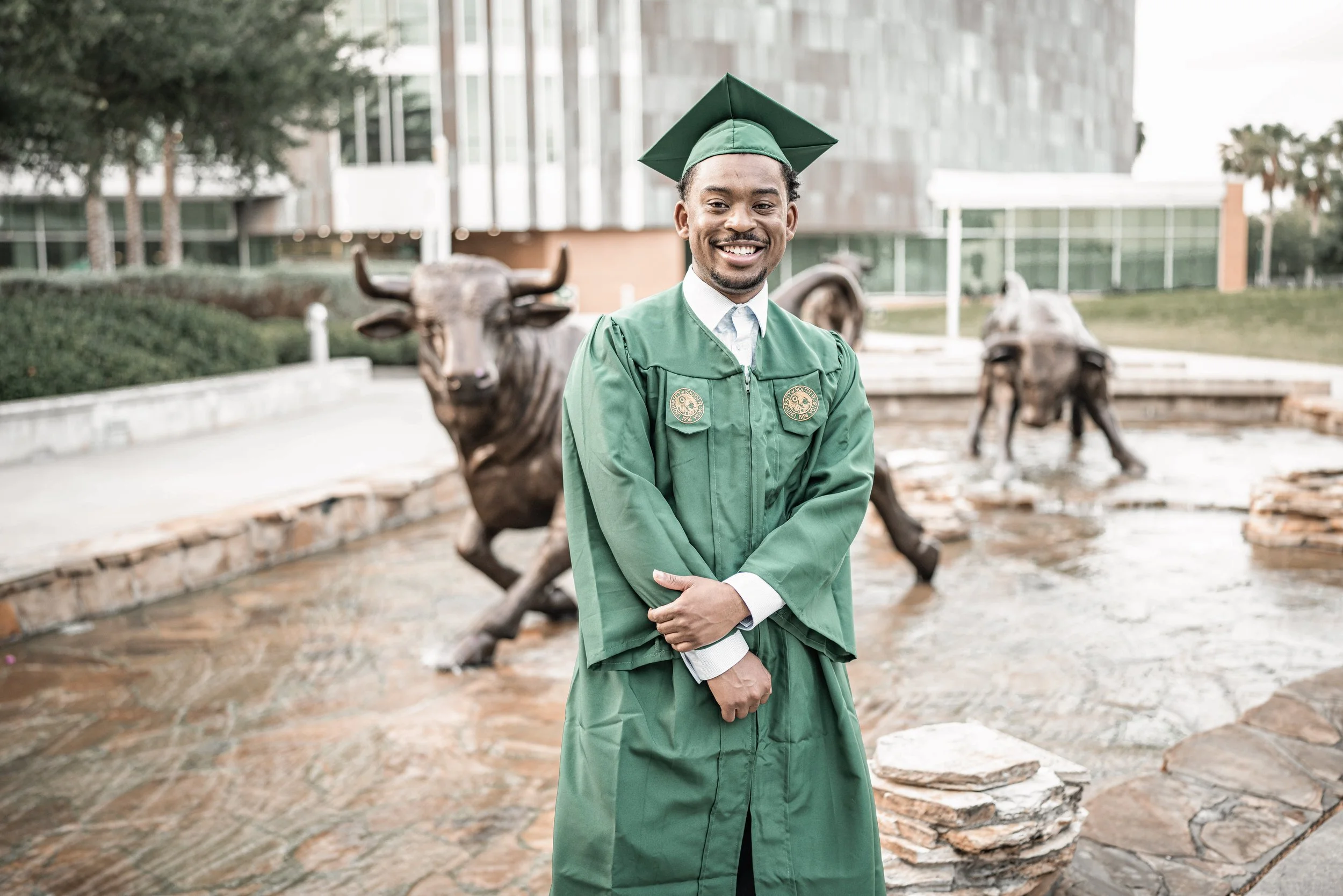 A smiling man in a green graduation gown and cap standing outdoors, with bronze bull statues and modern glass buildings in the background.