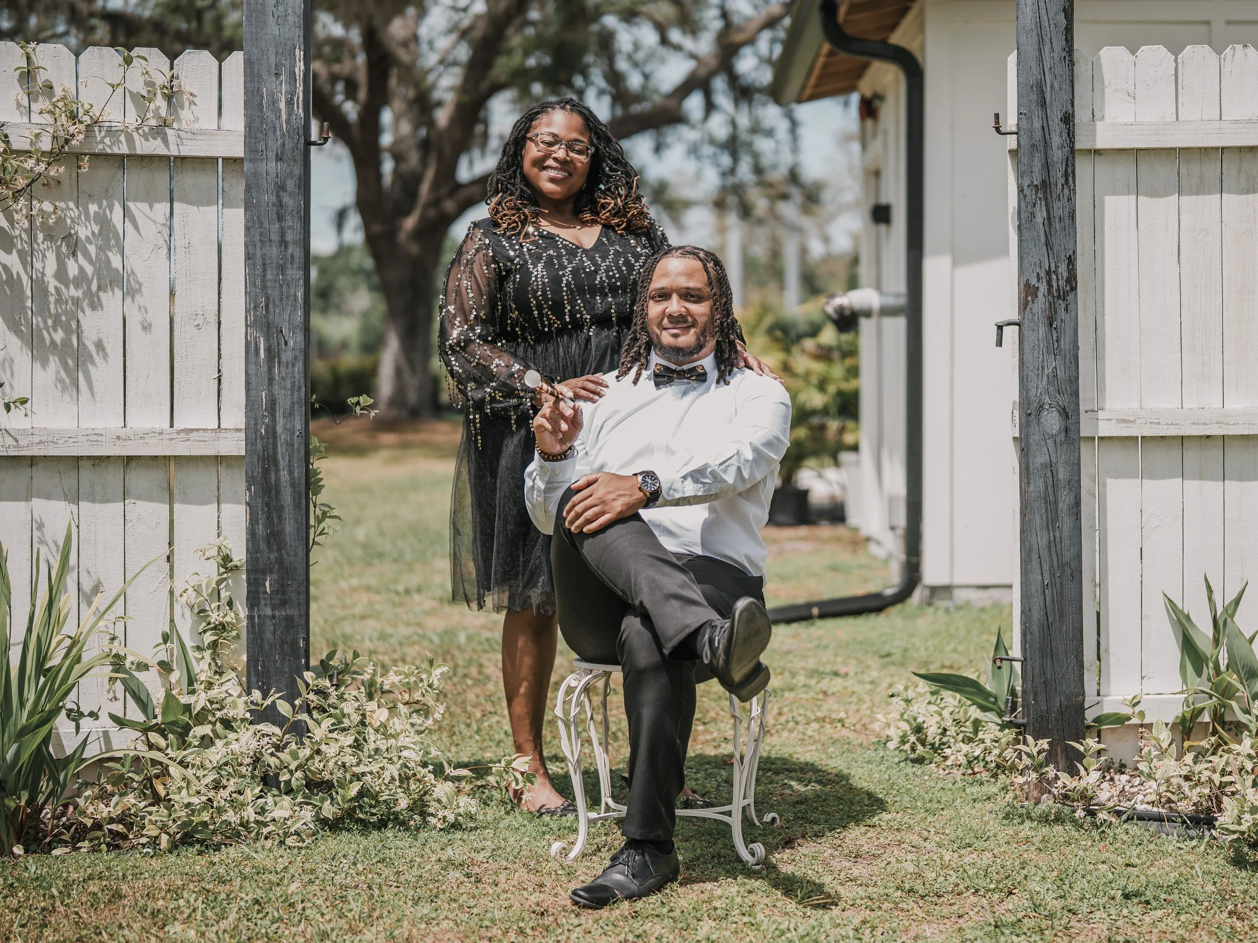 A woman standing behind a man sitting on a decorative metal chair, both smiling outdoors behind a white fence with greenery and trees in the background. The woman has black, curly hair and glasses, wearing a black dress with embellishments. The man h