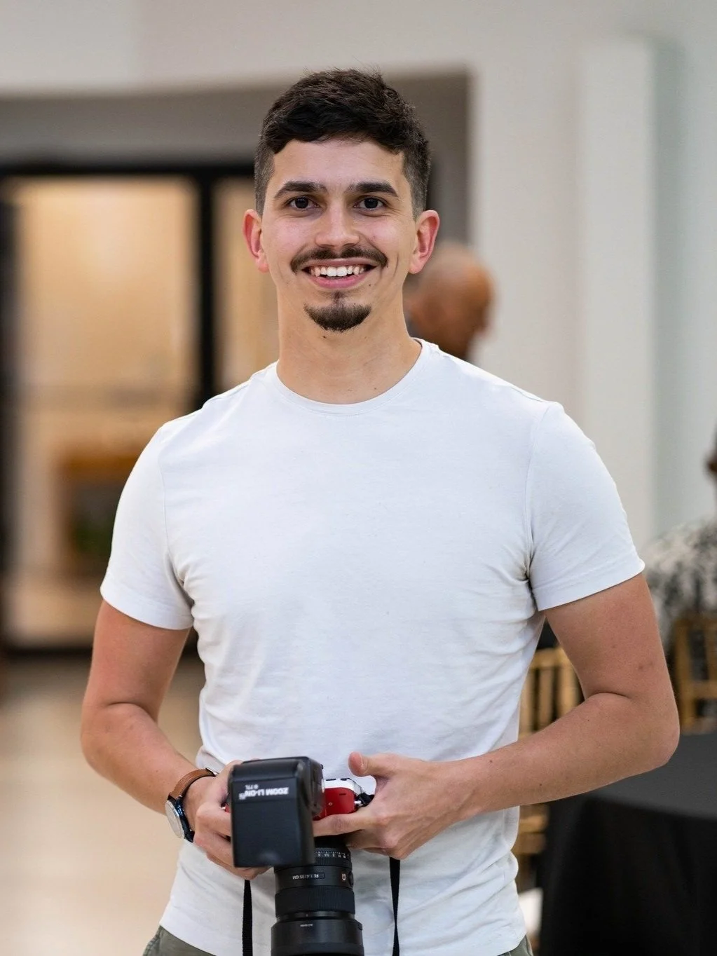 A young man with dark hair, facial hair, and a smile, holding a professional camera, standing indoors with a blurred background.