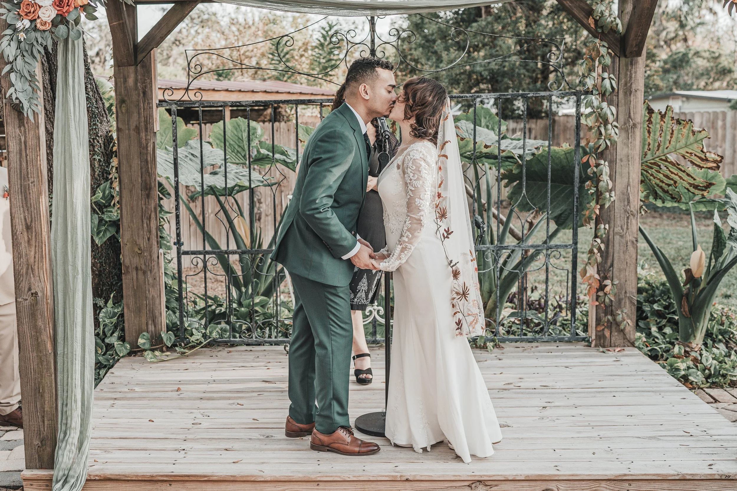 A wedding ceremony taking place outdoors on a wooden platform, with a bride and groom kissing. The groom is wearing a green suit, and the bride is in a white lace gown. Decorations include greenery and flowers, and a officiant is standing behind the 