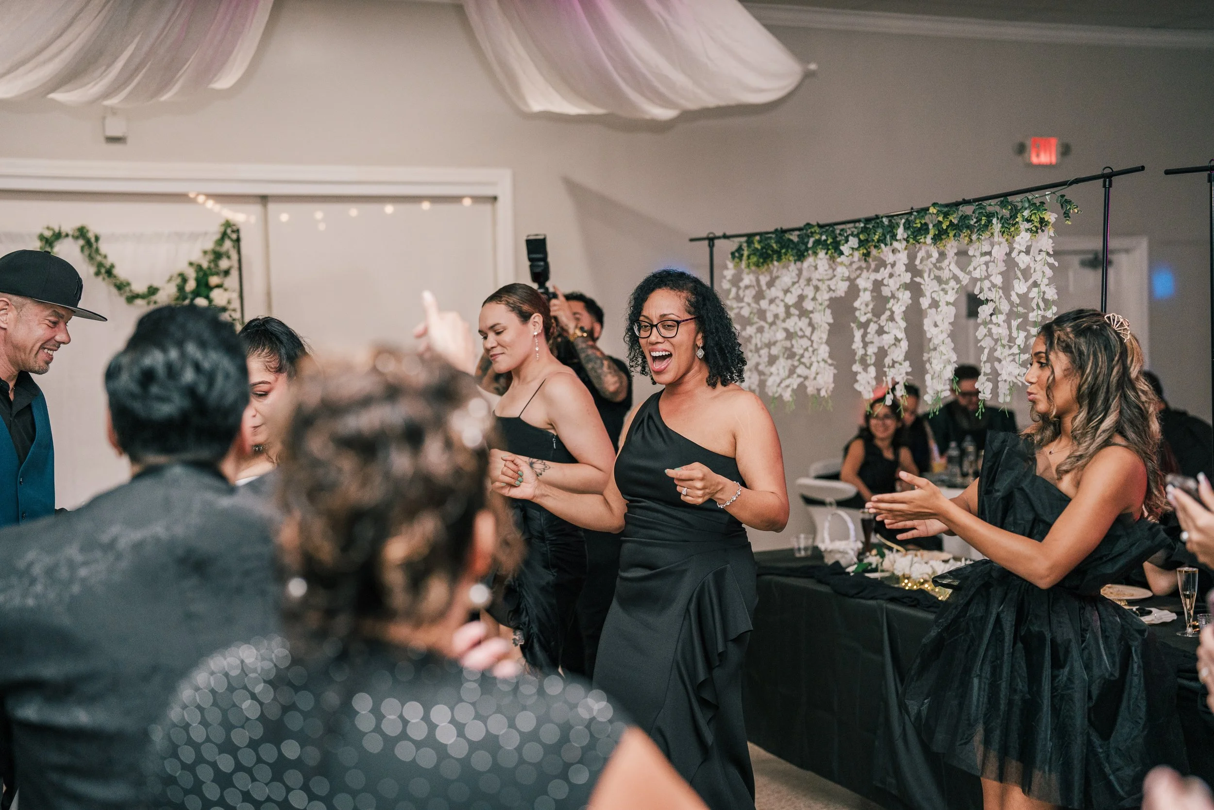People dance and socialize at a celebration or event, with floral decorations and a festive atmosphere.