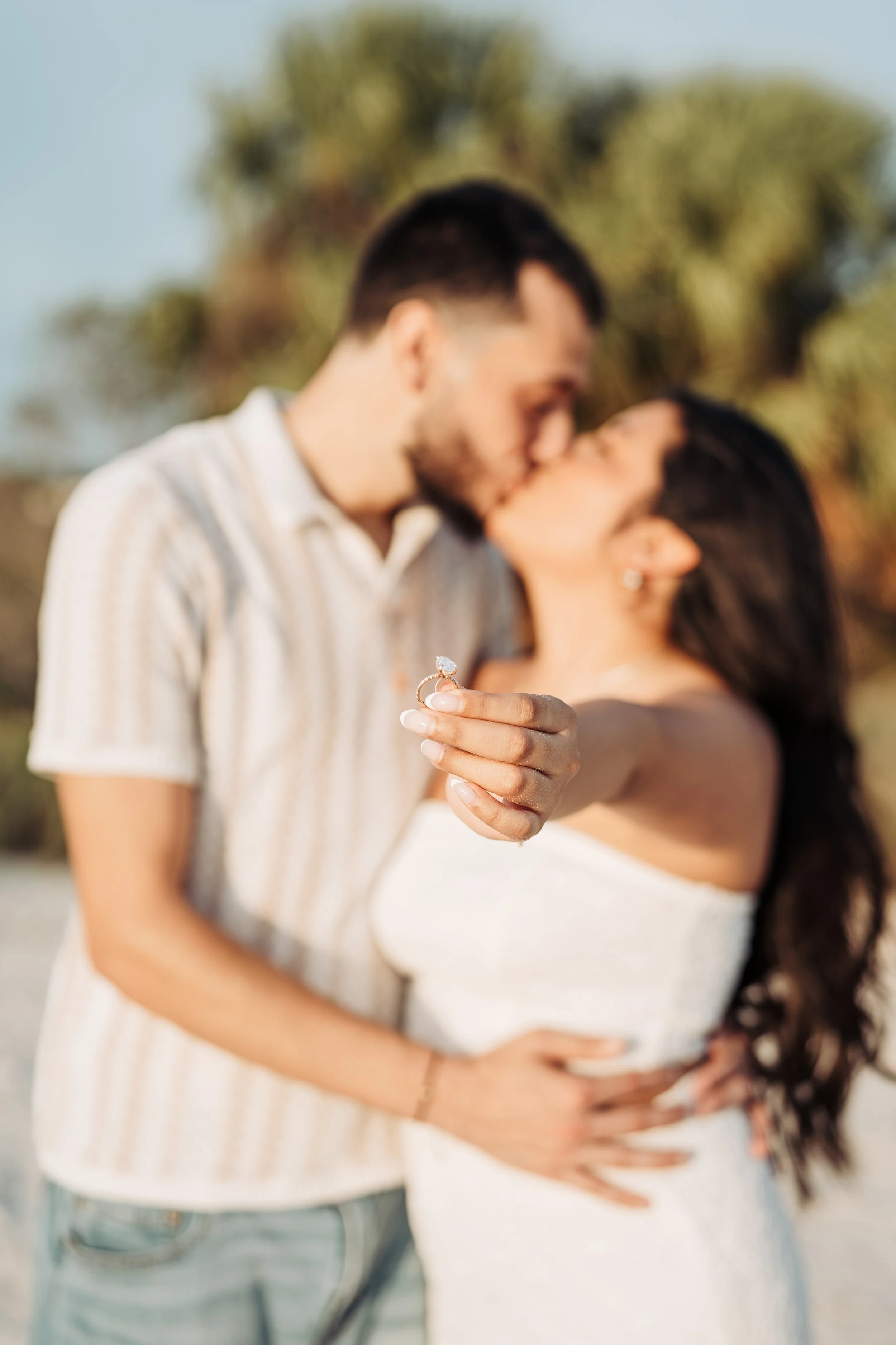 A couple engaging in a romantic moment outdoors, with the man kissing the woman on her forehead while she holds up a ring in her hand.