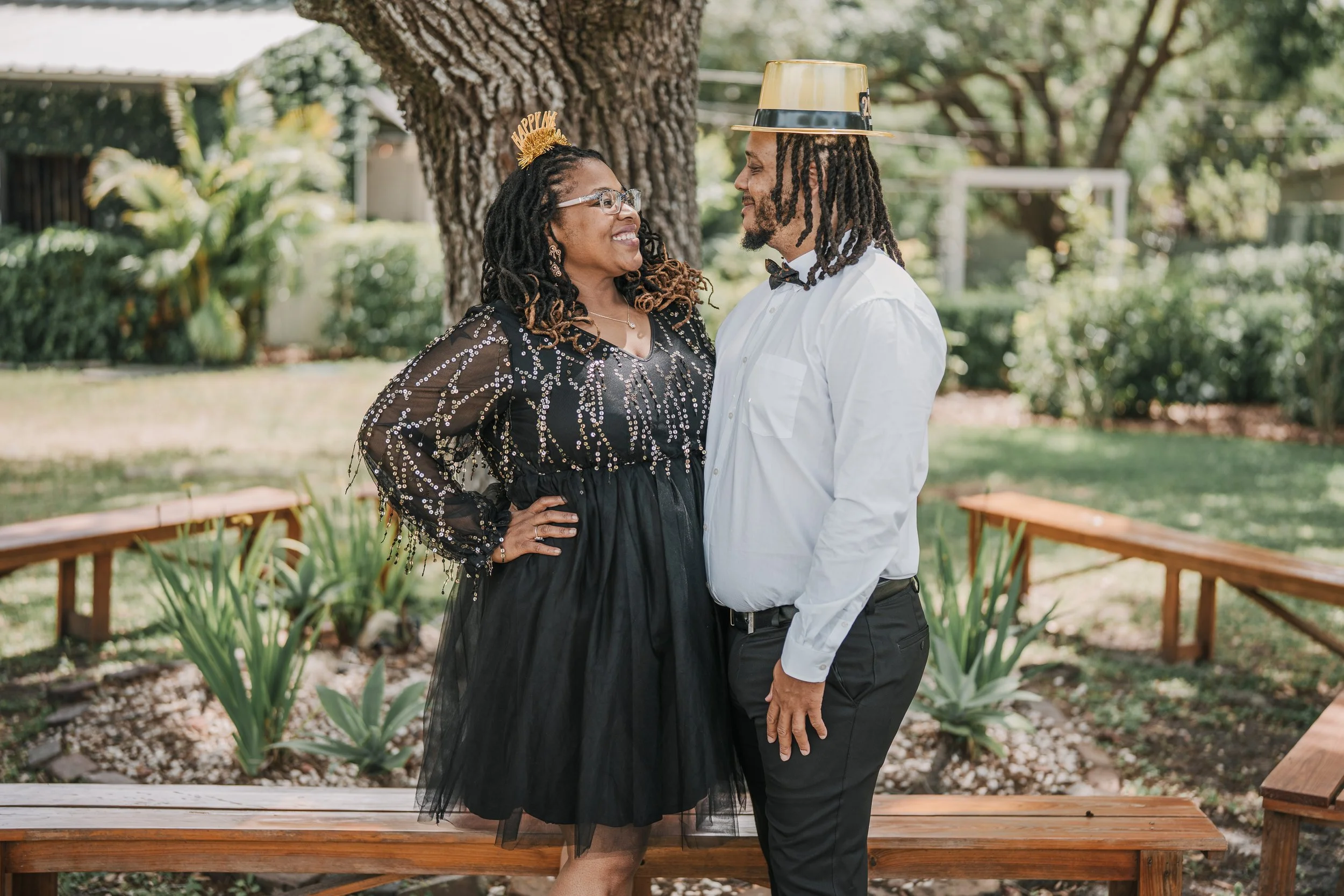 A woman and man celebrating an event outdoors near a large tree, with the woman wearing a 'Happy' headband and the man wearing a party hat, smiling and looking at each other.