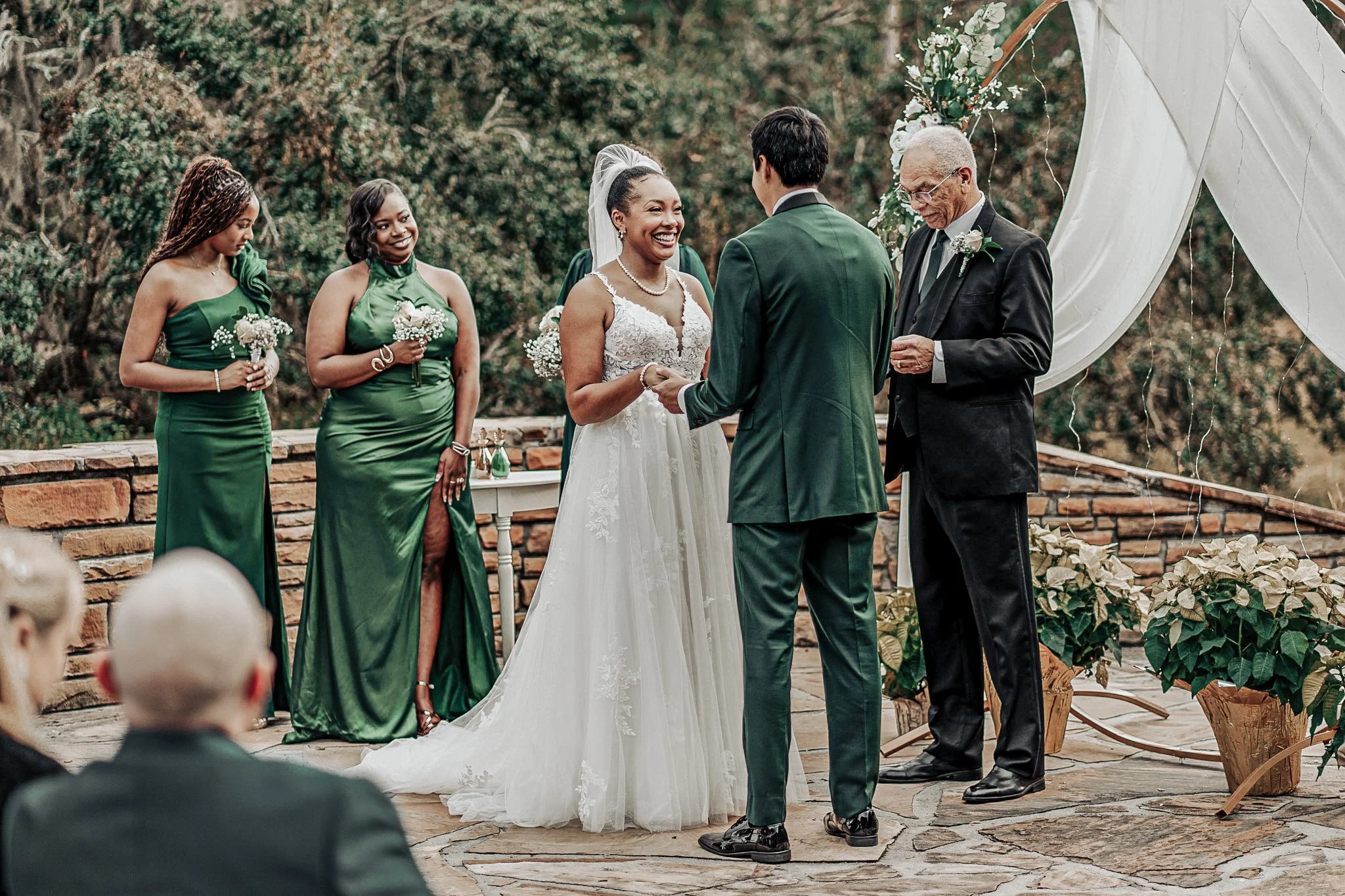 A wedding ceremony outdoors with a bride and groom holding hands and smiling, surrounded by bridesmaids and an officiant, with trees and decorations in the background.