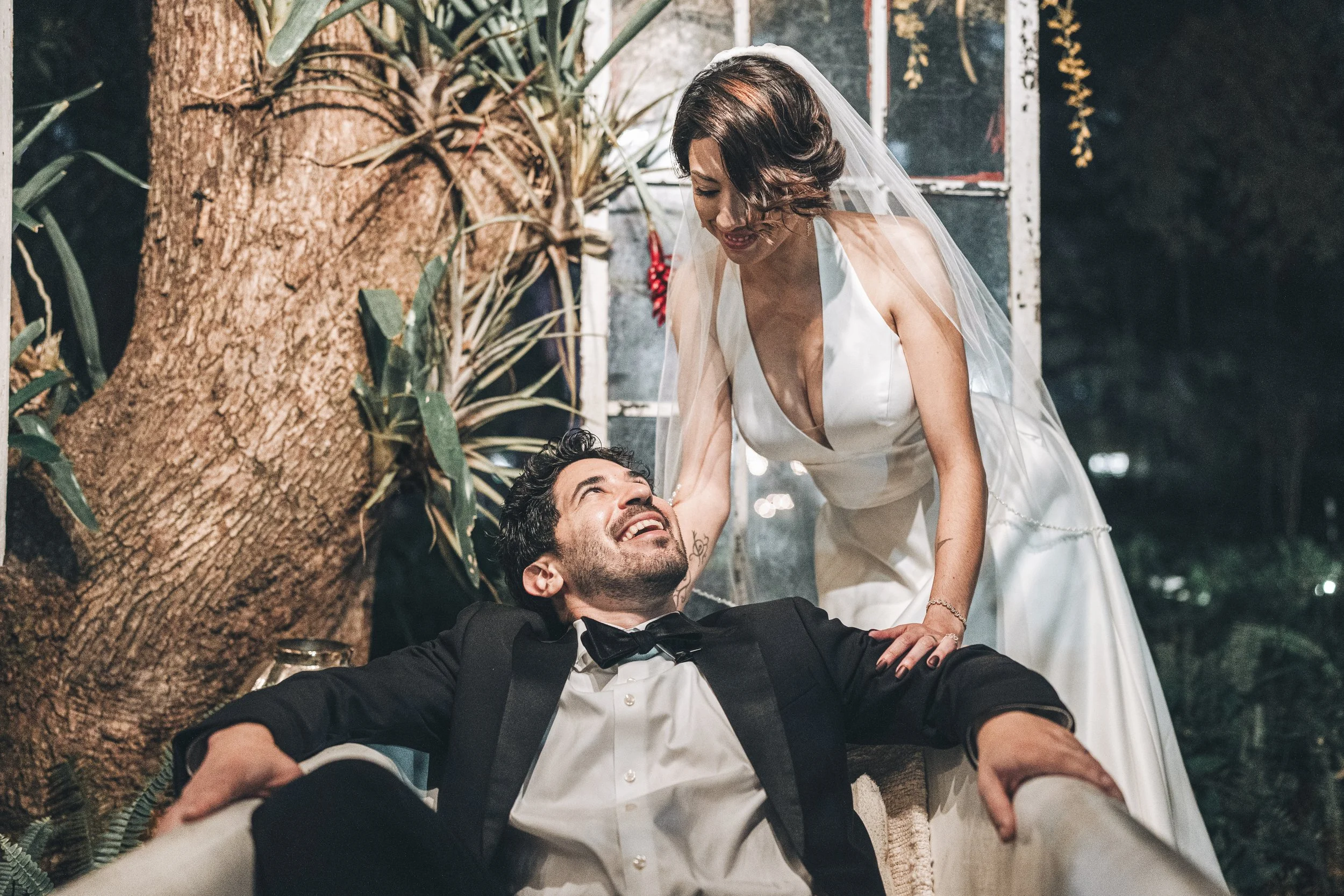A bride and groom enjoying a moment together outdoors, with the bride leaning over the groom who is lying on the ground, surrounded by greenery and trees.