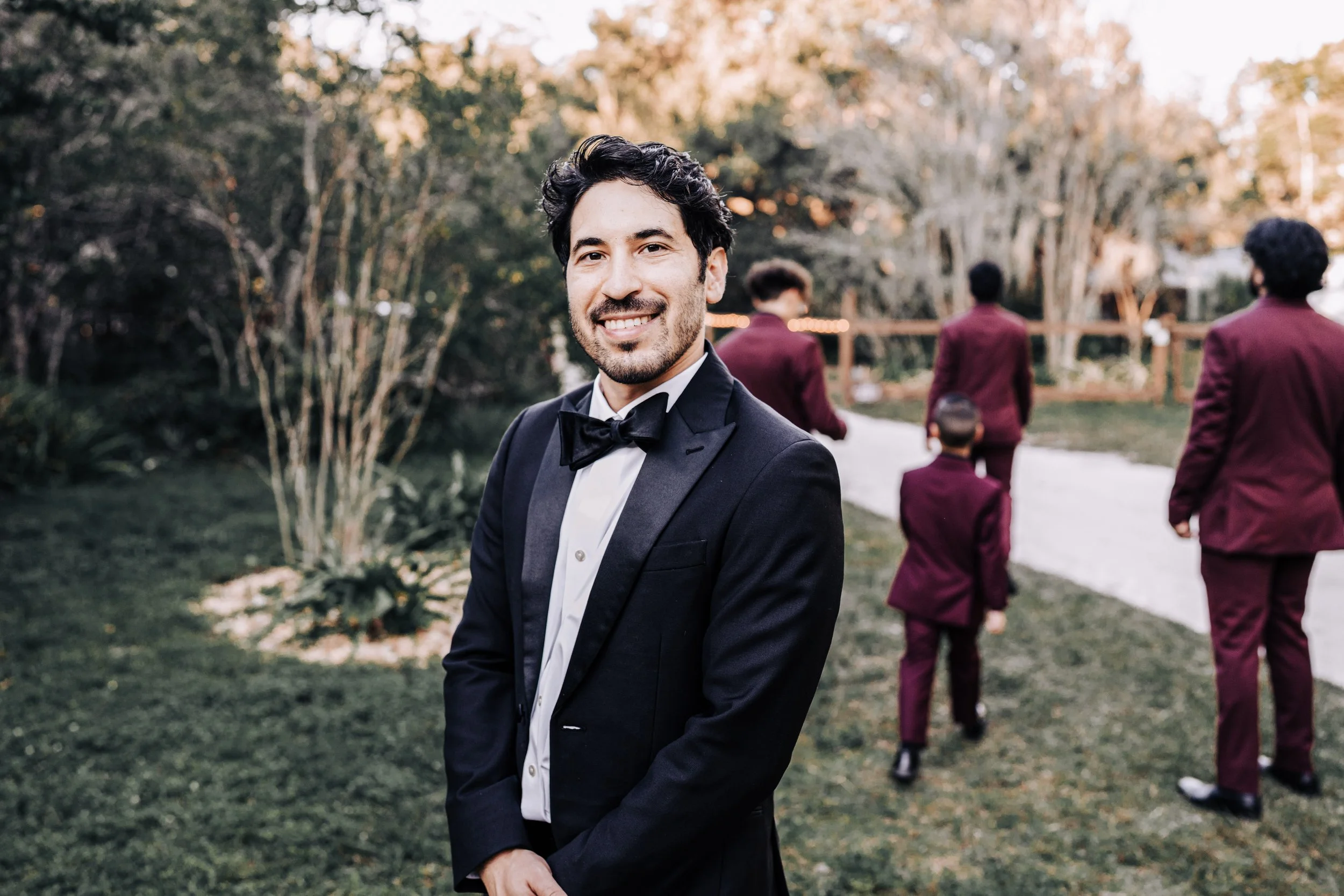 A man in a black tuxedo with a bow tie smiling at the camera outdoors, with people in maroon suits walking away in the background near trees and greenery.
