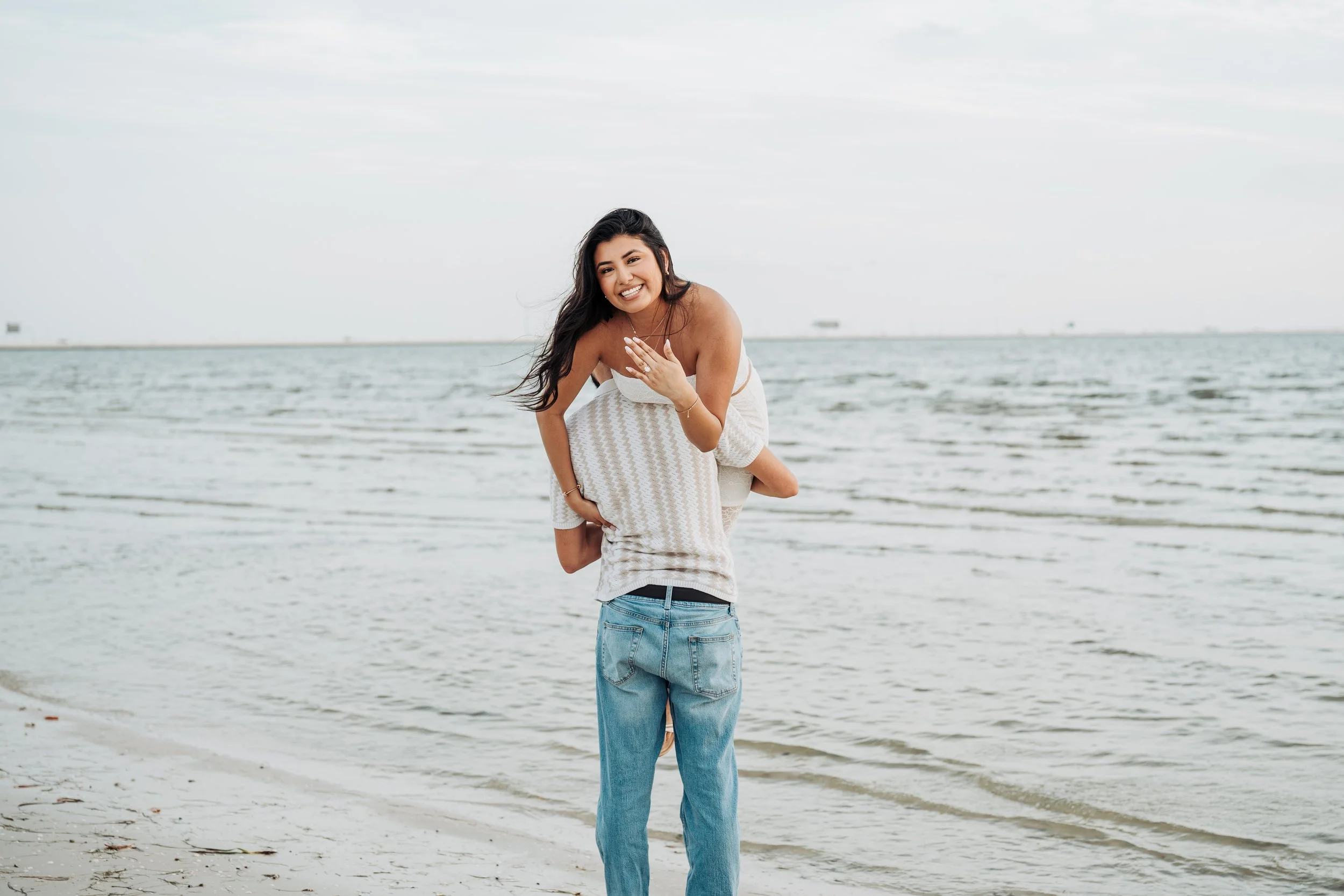 A young woman with long dark hair smiling while being carried piggyback on someone at the beach.
