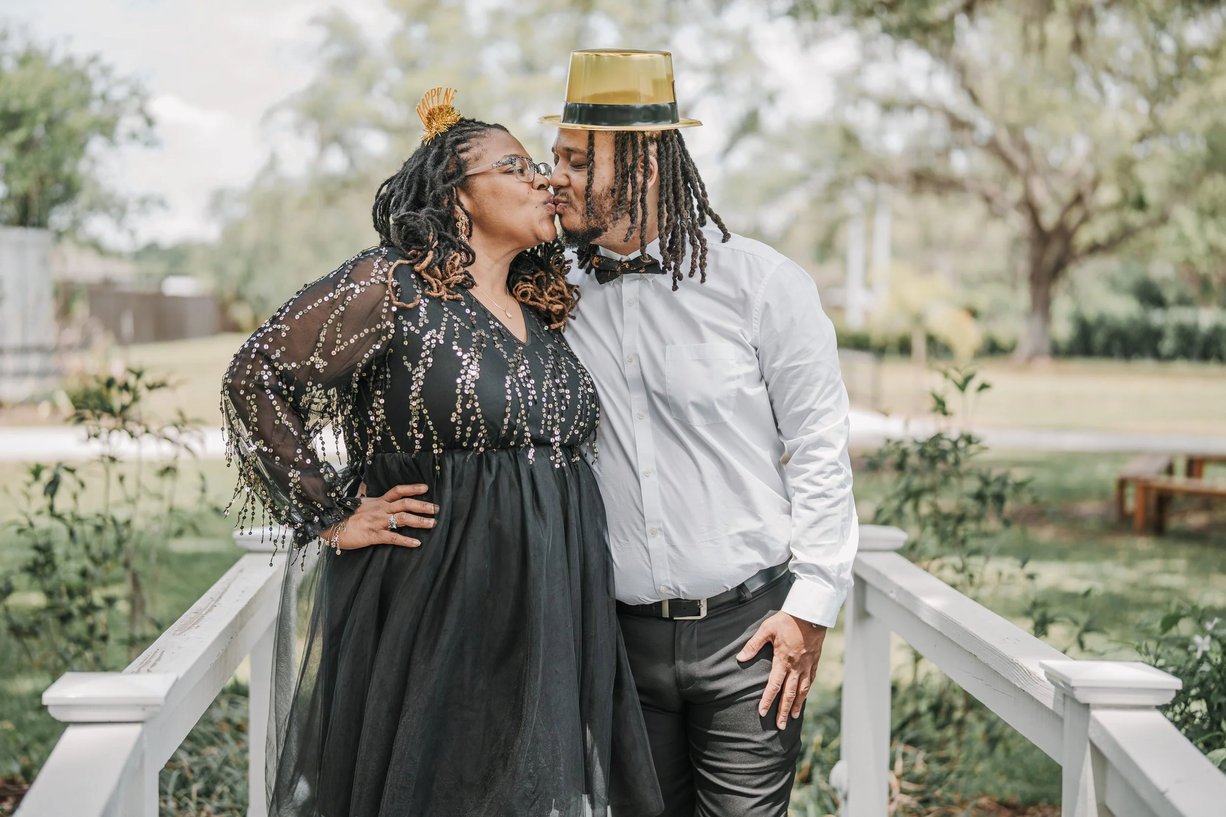 A couple standing on a white bridge outdoors, about to kiss, with the woman wearing a black sequined dress, glasses, and a 'Happy New Year' headband, and the man wearing a white shirt, black pants, a bow tie, and a golden top hat.