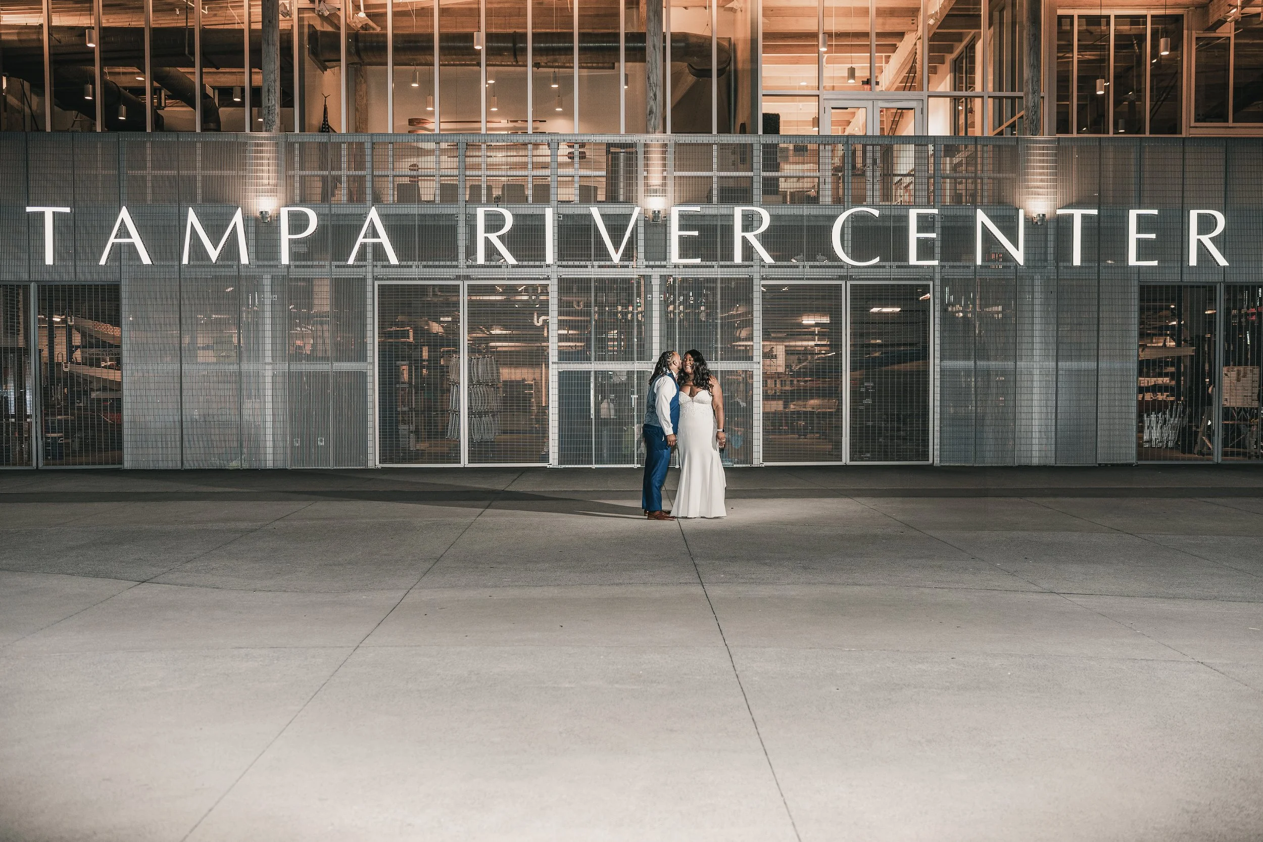 A couple dressed in wedding attire standing under a large sign that reads 'TAMPA RIVER CENTER.' The building has a modern industrial design with glass walls, exposed metal framework, and interior lighting visible through the windows.