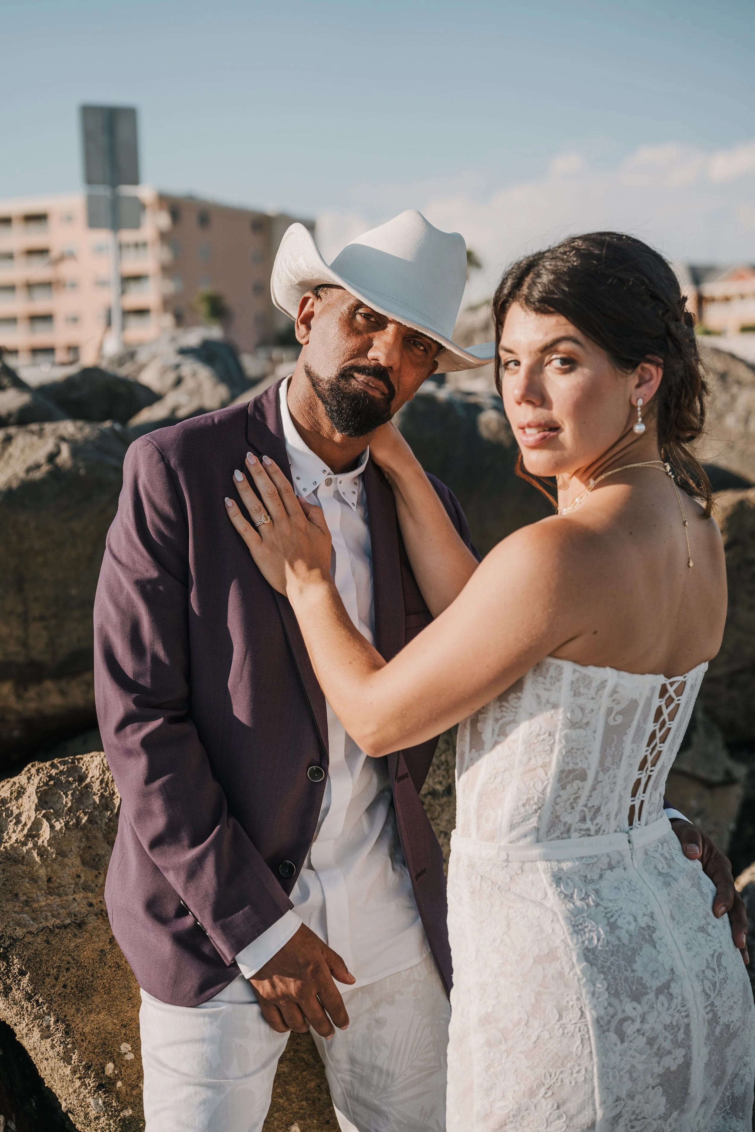 A man wearing a white cowboy hat, a dark blazer, and white pants, and a woman in a white lace dress with pearl earrings, embracing outdoors on a sunny day with rocks and a building in the background.