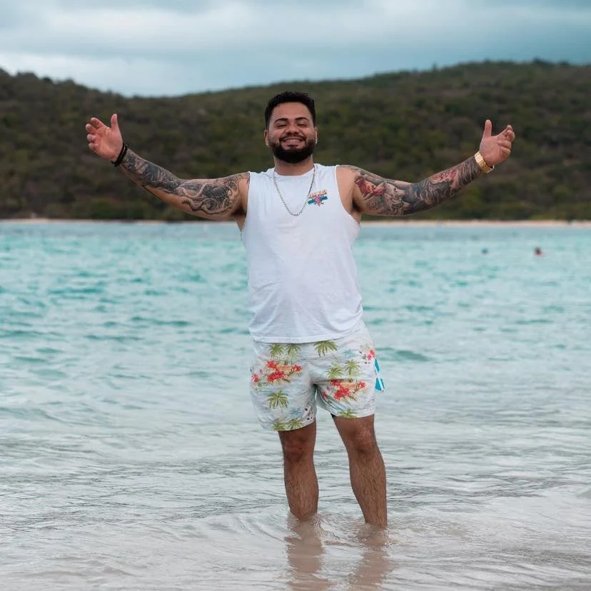 A man standing in the ocean water with arms outstretched and smiling, wearing a white sleeveless shirt and tropical-patterned shorts, with a background of a hilly landscape and cloudy sky.