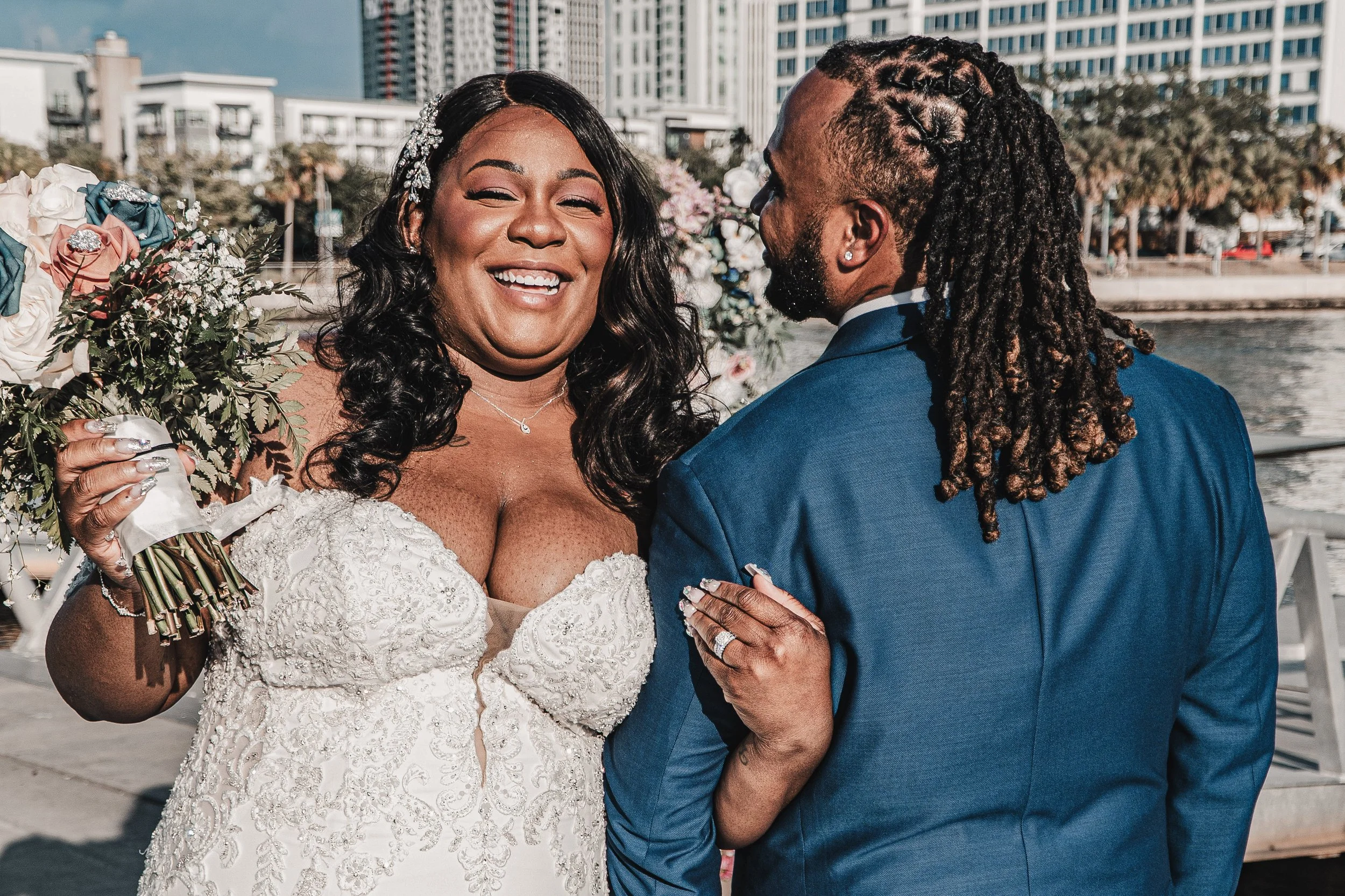 A joyful bride and groom share a close moment during their wedding celebration near a body of water with city buildings in the background. The bride wears a white lace gown and the groom is in a blue suit with dreadlocks.