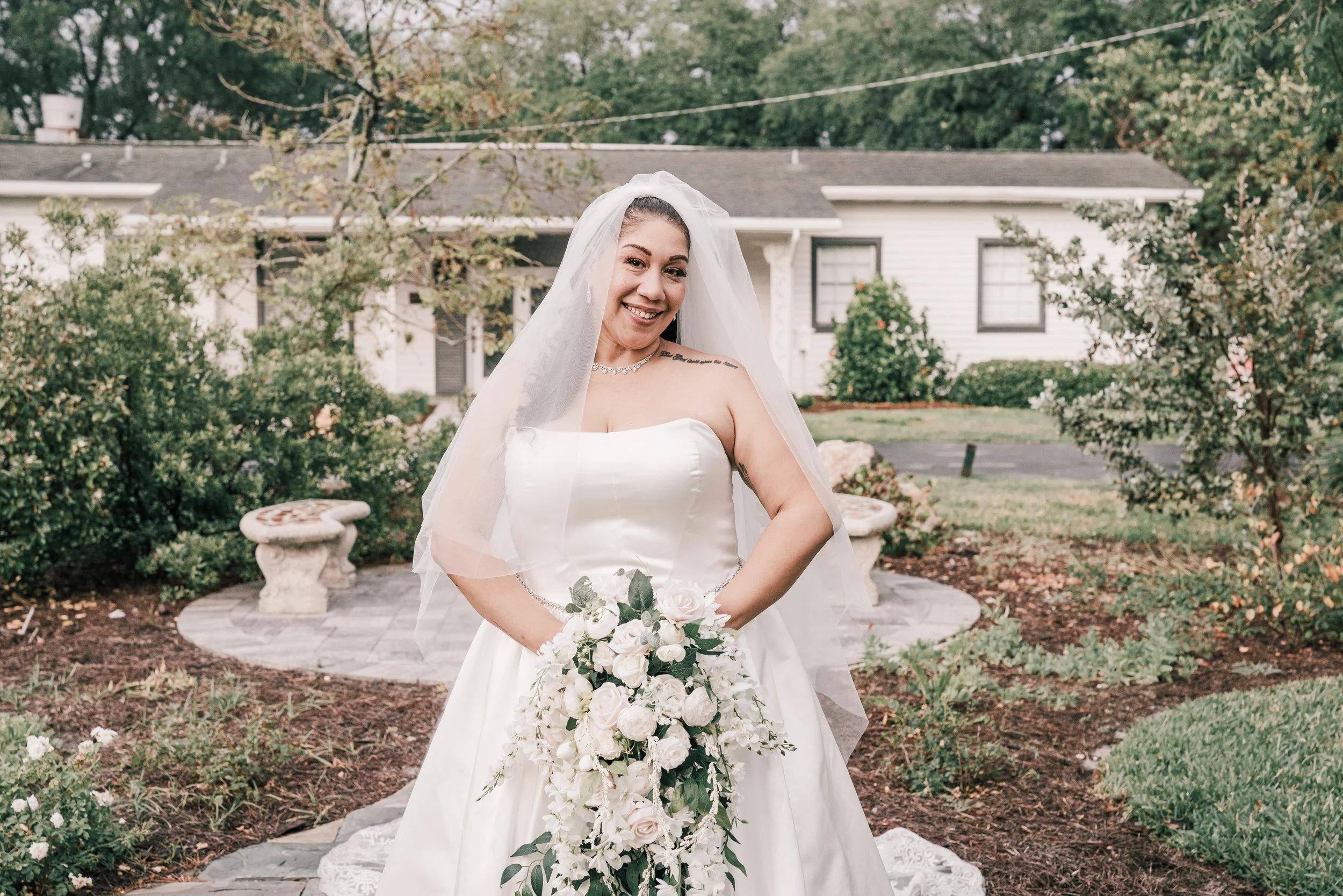 A bride smiling outdoors in a white wedding gown and veil, holding a bouquet of white and pale pink roses, standing in front of a garden with greenery, stone benches, and a house in the background.