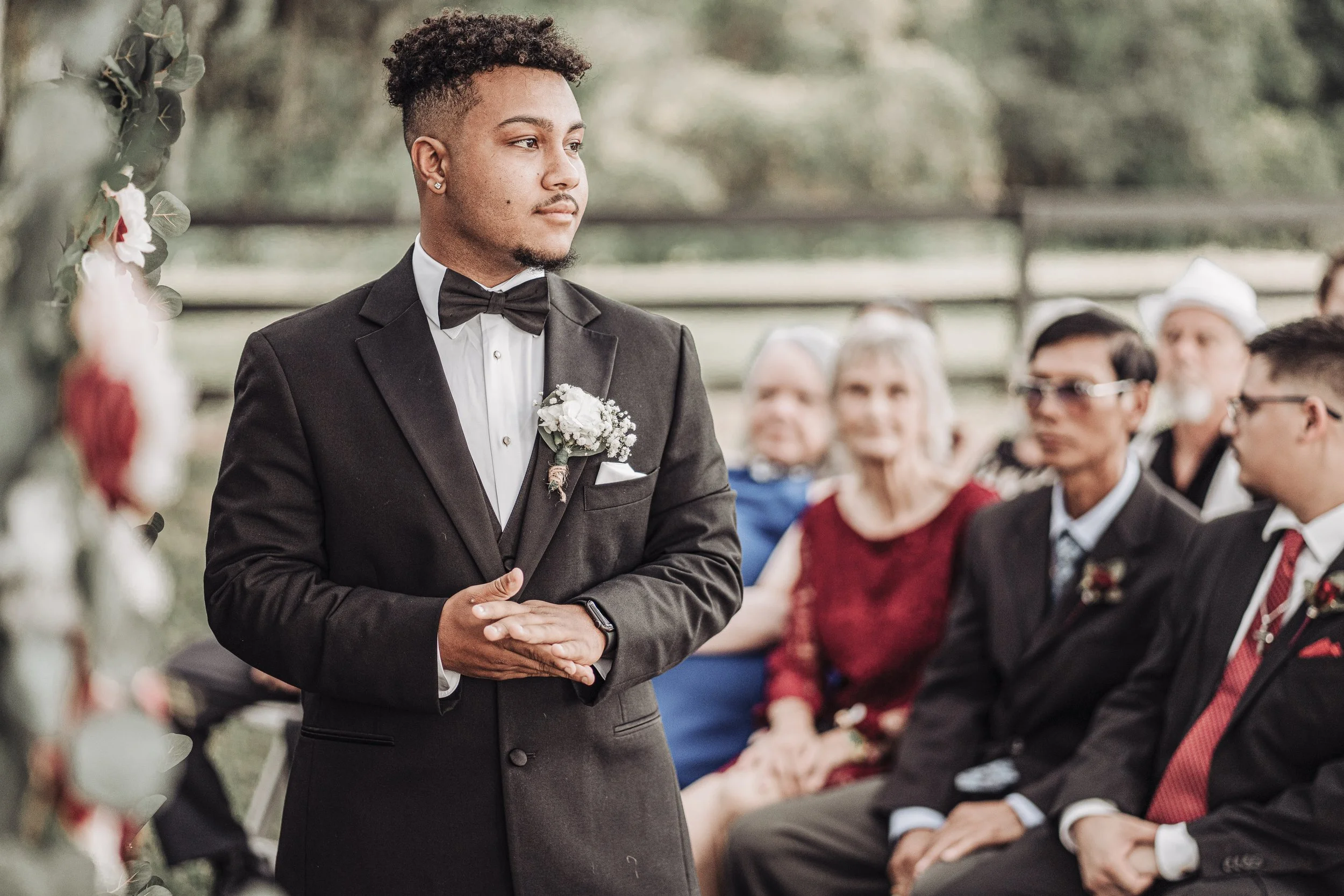 A groom in a black tuxedo with a bow tie and boutonniere standing during a wedding ceremony outdoors, with wedding guests sitting behind him.