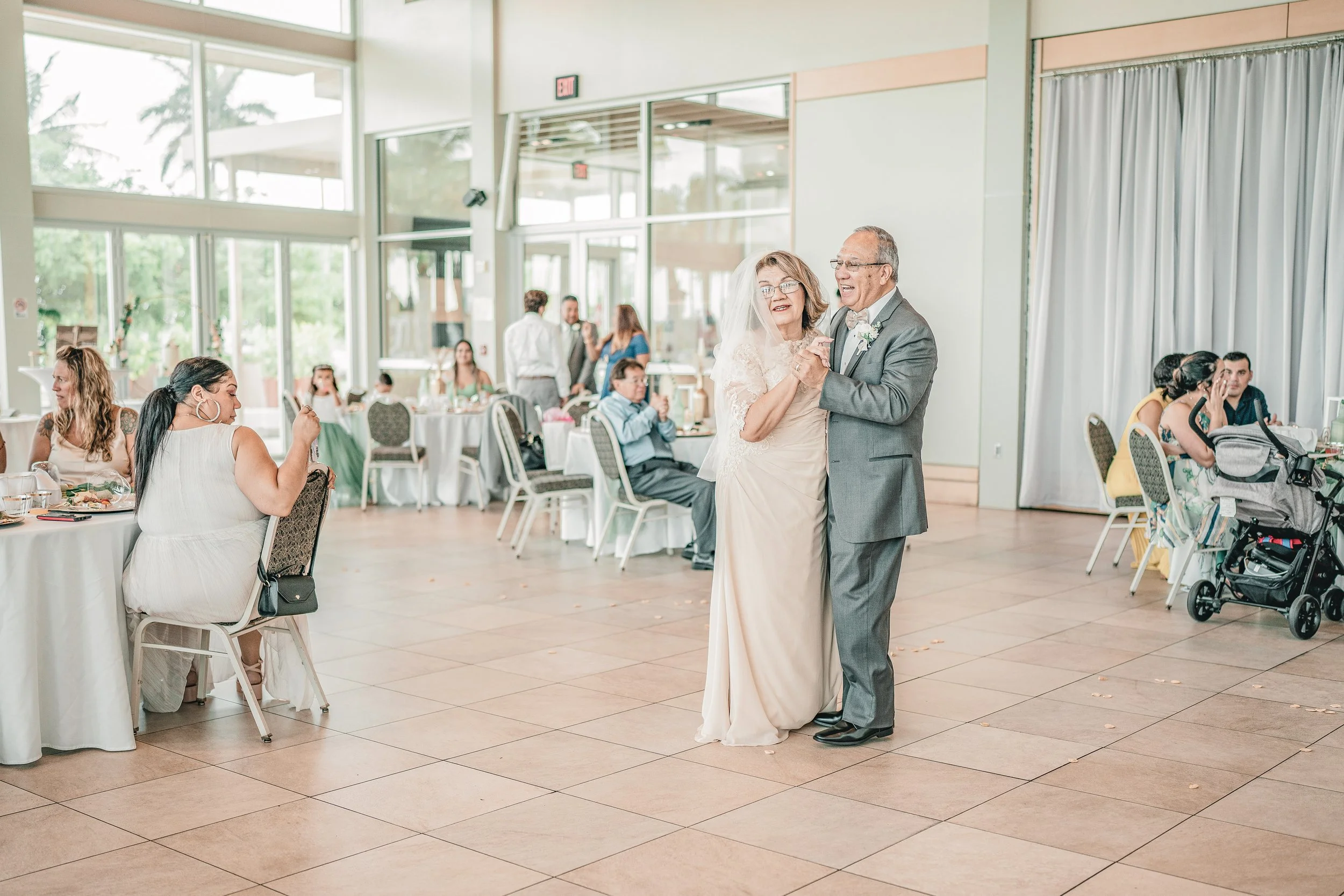 A senior couple dancing at a wedding reception in a bright, modern hall with large windows, while guests sit and watch at tables.