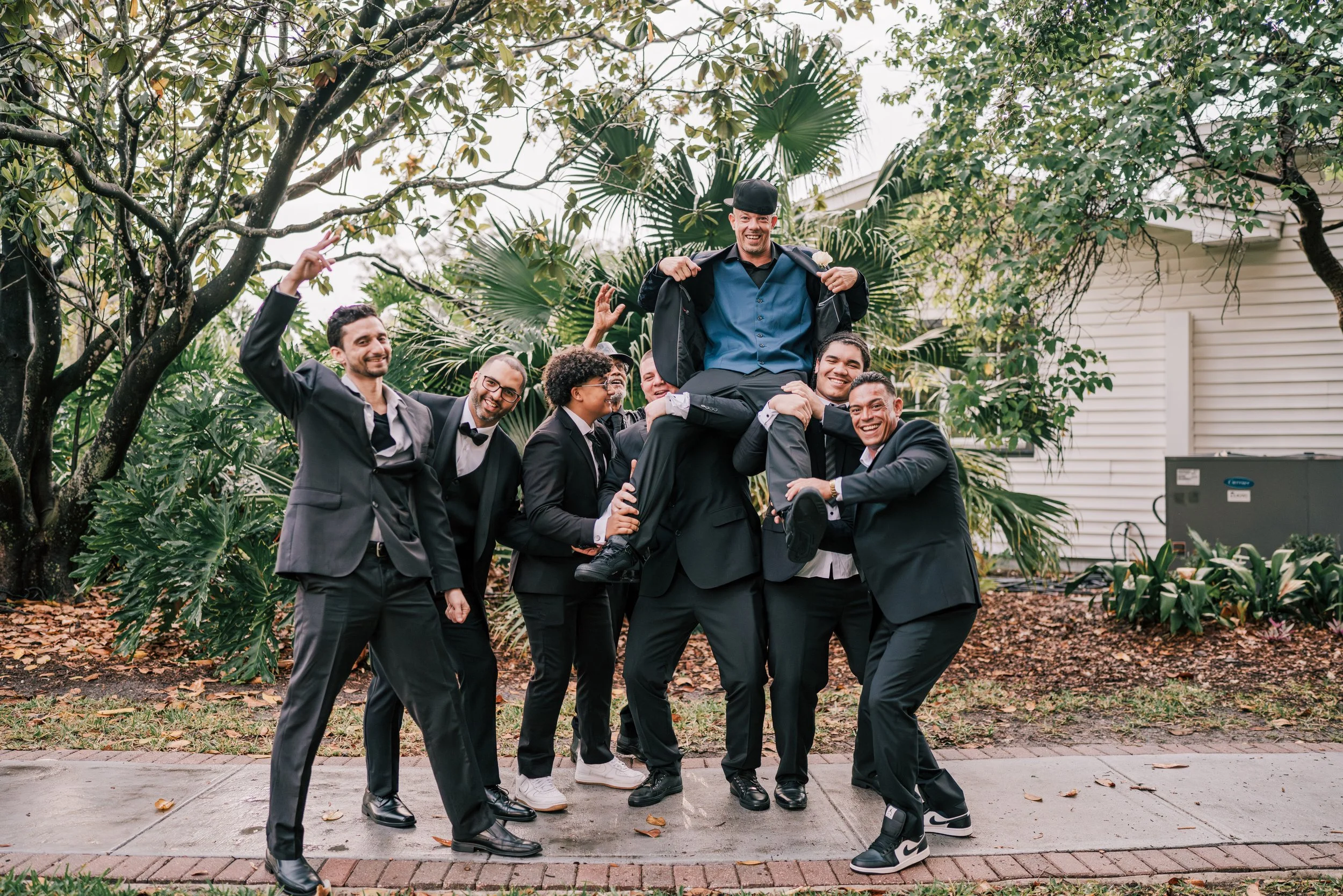 A group of seven men dressed in suits celebrating outdoors, with one man sitting on the shoulders of others, all smiling and appearing happy.