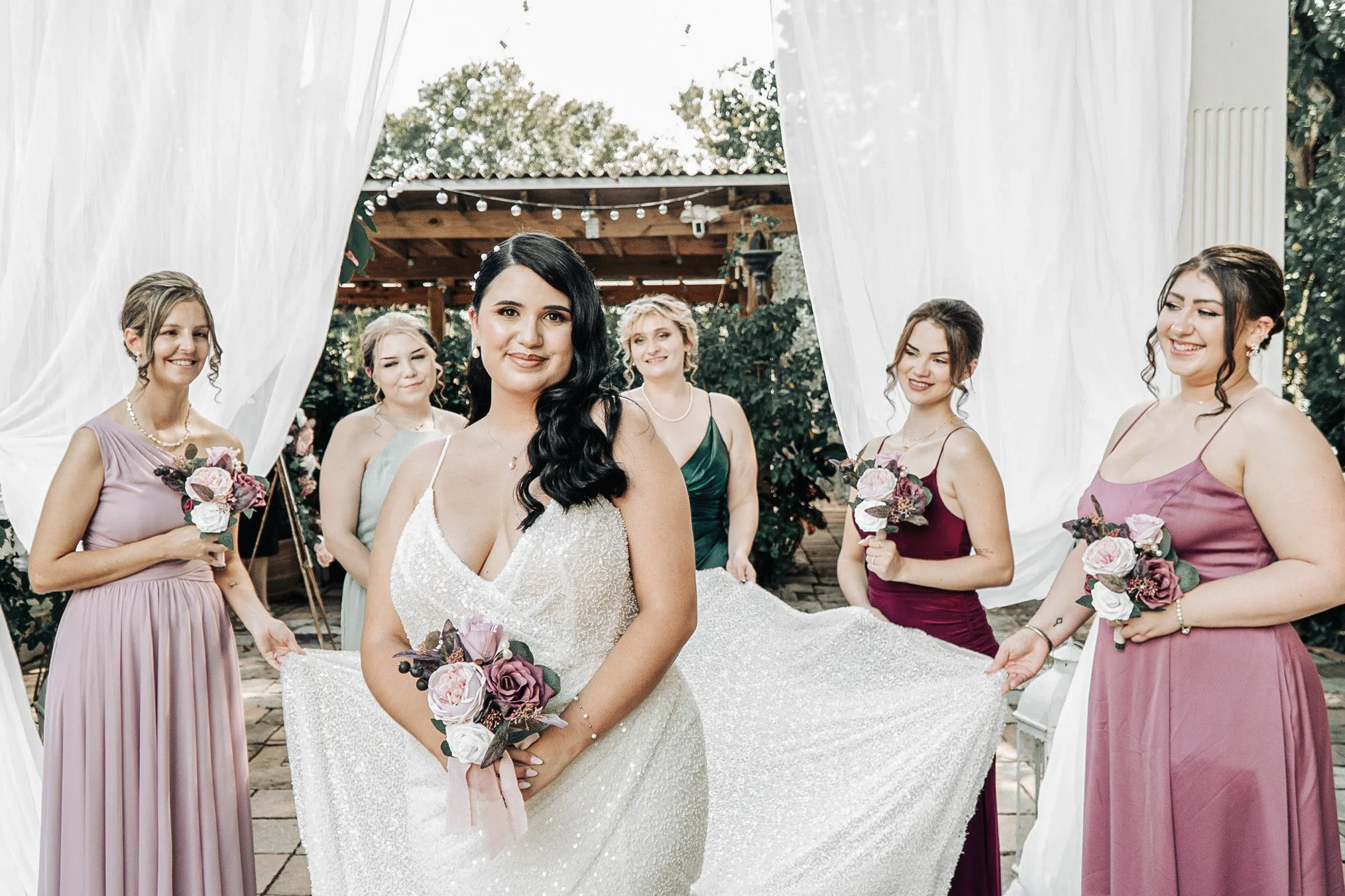 A bride in a white dress, smiling, holding a bouquet, surrounded by bridesmaids dressed in pastel and burgundy, at an outdoor wedding with a decorated backdrop.