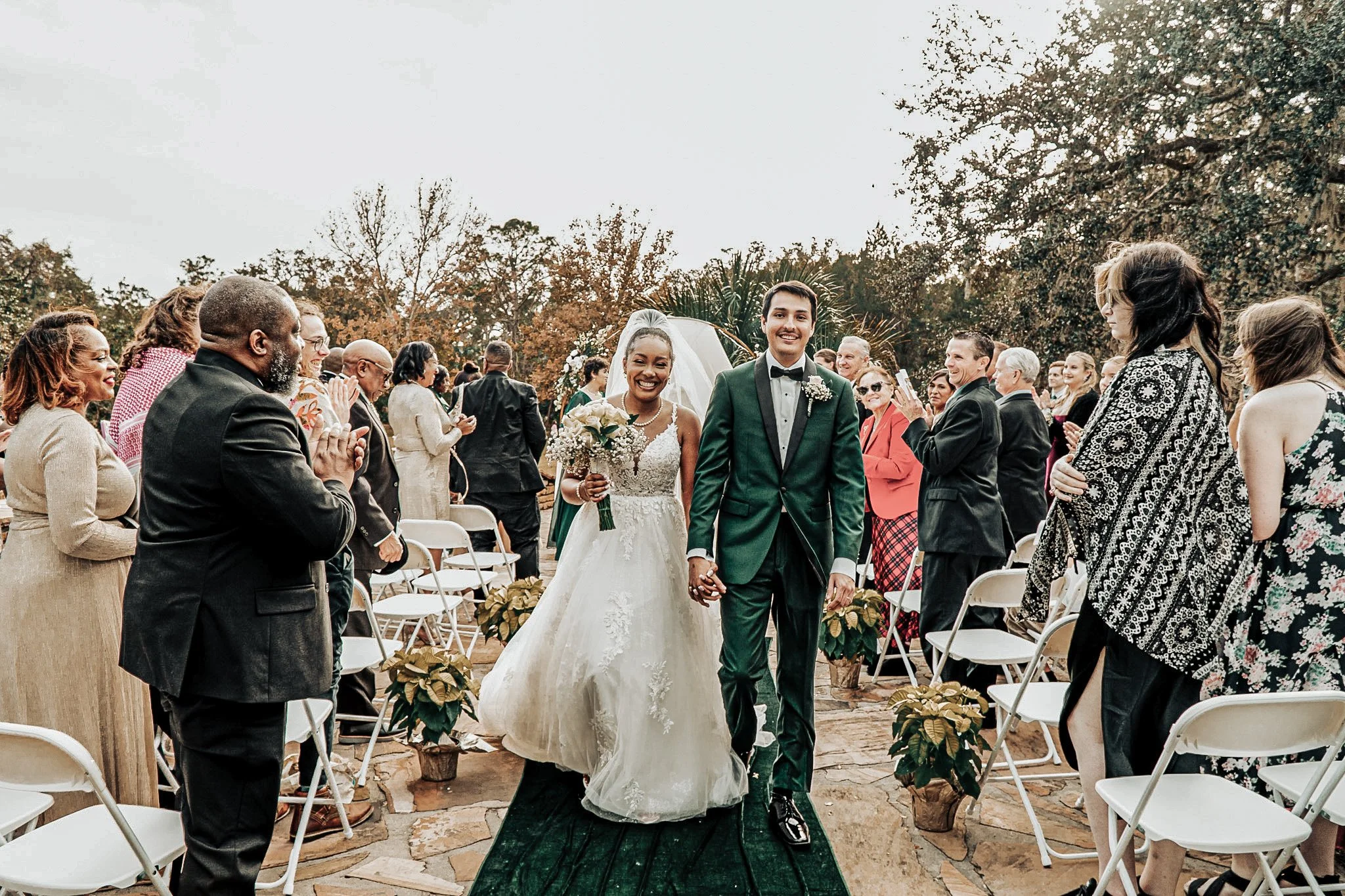 A bride and groom walking down the aisle after their wedding ceremony, holding hands and smiling, surrounded by seated guests clapping and taking photos, outdoors with trees in the background.