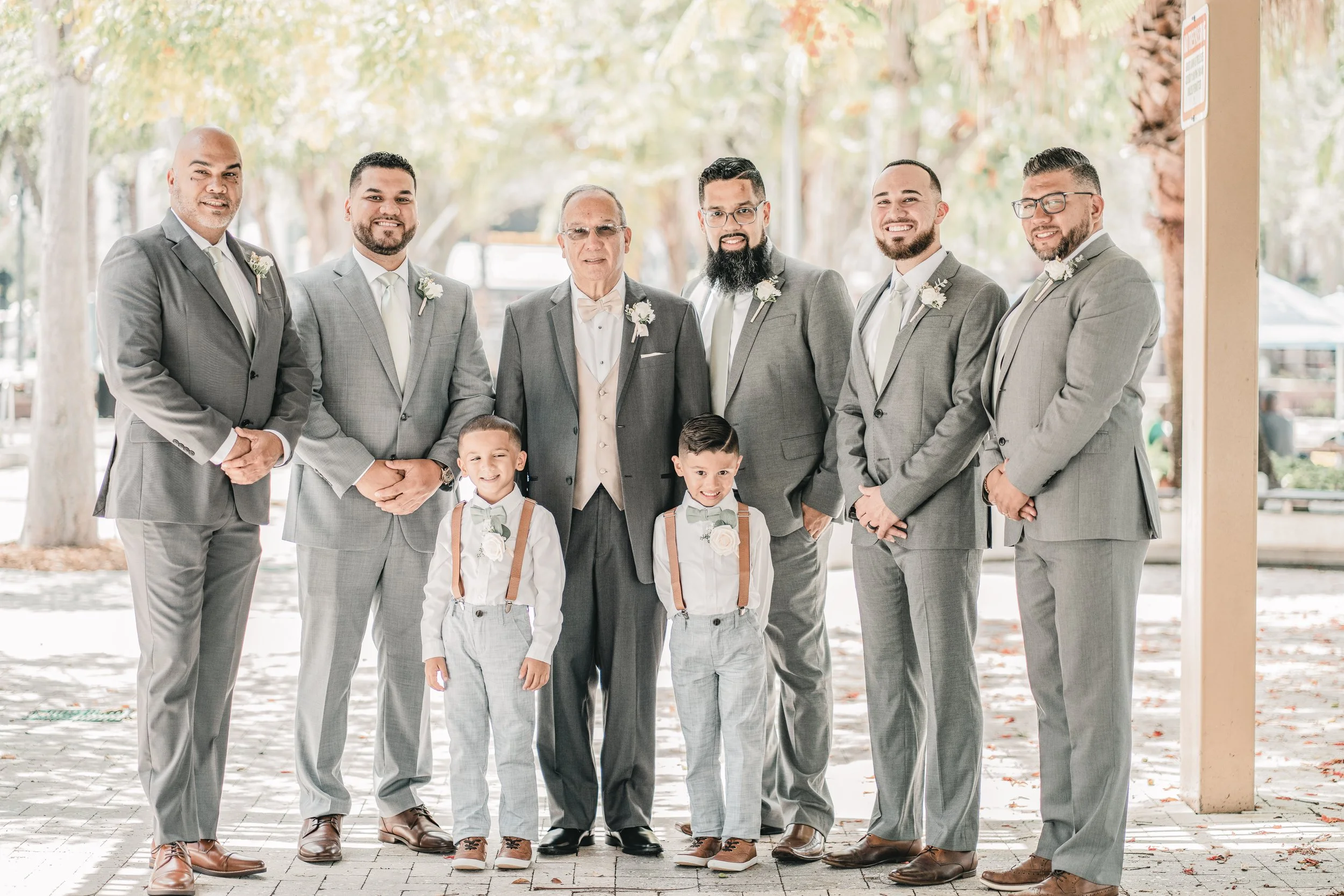 A group of seven men and two boys dressed in suits, standing outdoors with trees and fall foliage in the background, smiling at the camera.