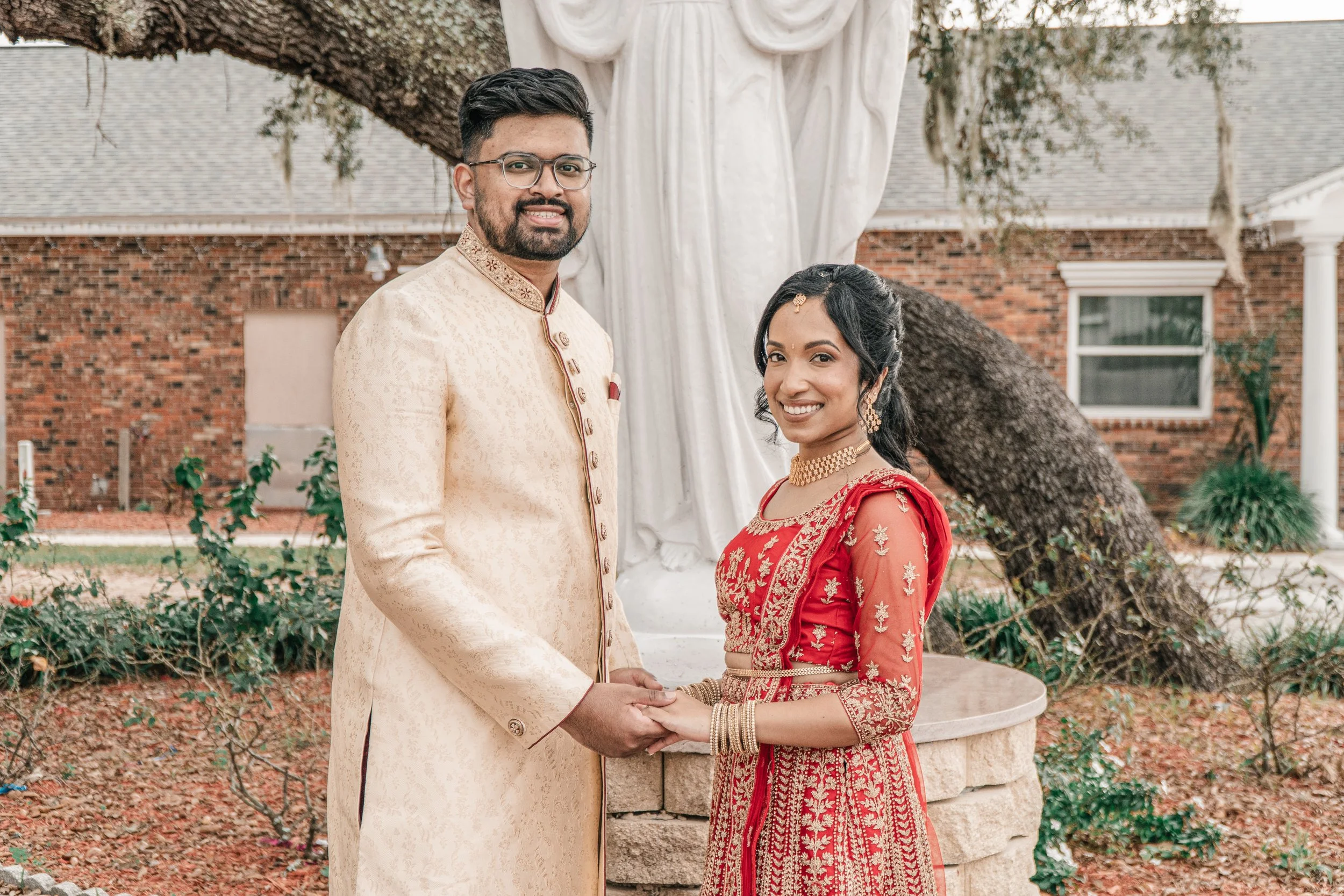 A couple dressed in traditional wedding attire holding hands outdoors in front of a large outdoor sculpture of a woman draped in fabric. The man wears a cream-colored sherwani with embroidery, and the woman wears a red sari with gold embroidery and j