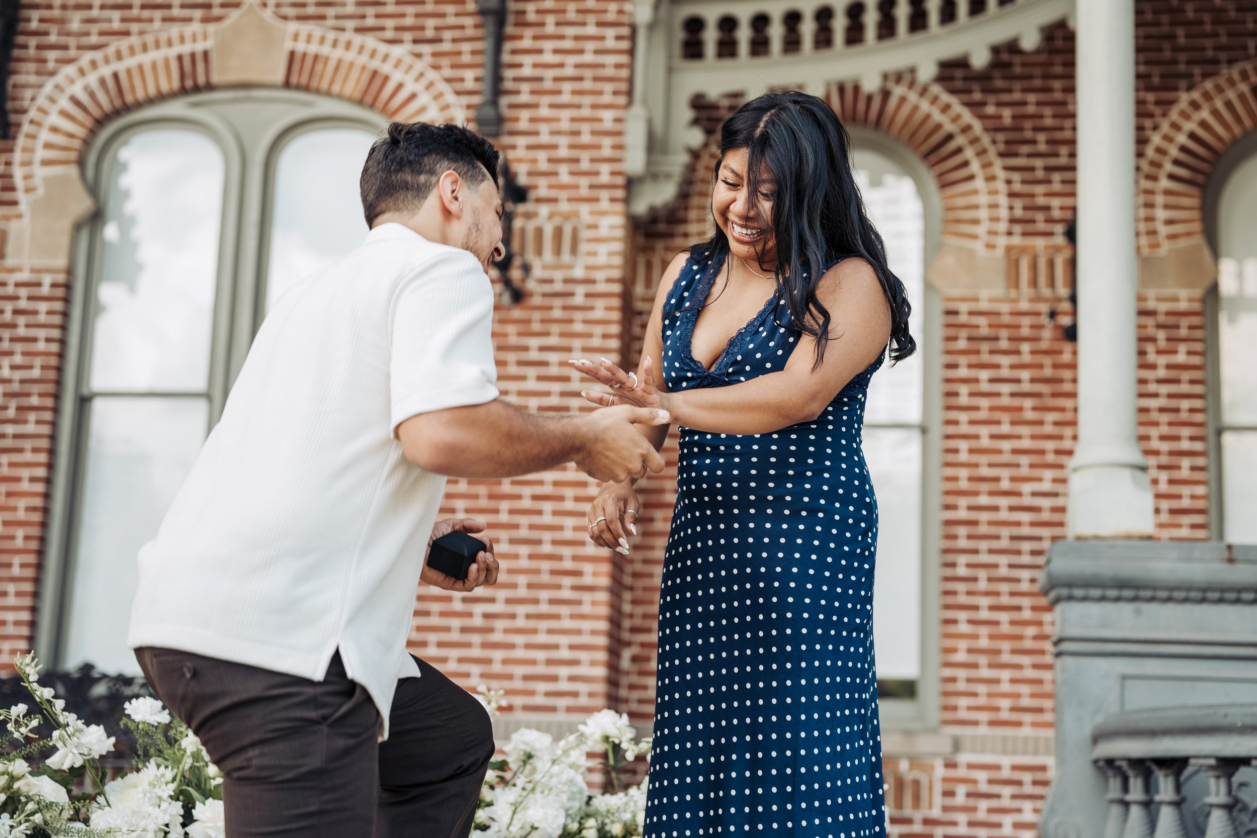 A woman in a blue polka dot dress is smiling as she looks at a man proposing marriage. The man is kneeling, holding a small black box, with a brick building and white flowers in the background.