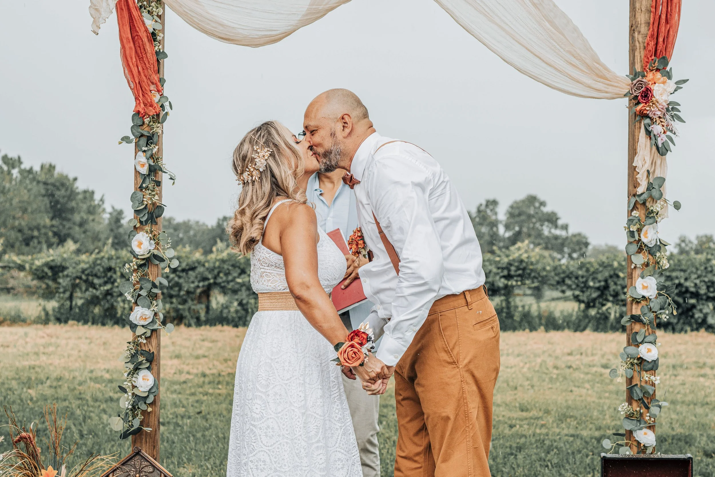 A couple getting married outdoors, holding hands and sharing a kiss under a decorated arch with flowers and fabric, with a person officiating in the background.