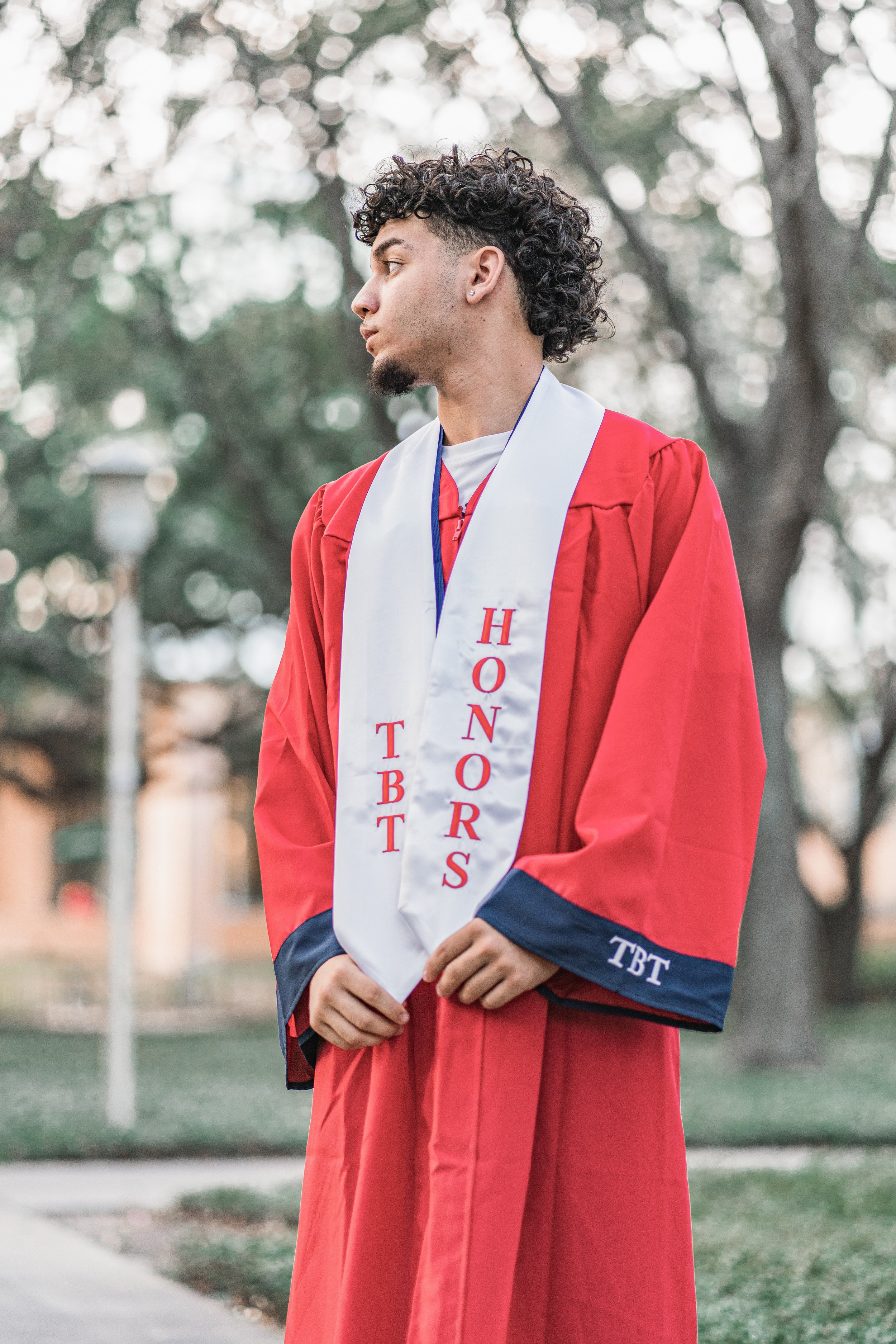 A young man in a graduation cap and gown standing outdoors with trees in the background. He is wearing a white stole with red text reading 'HONORS' and 'TBT'.