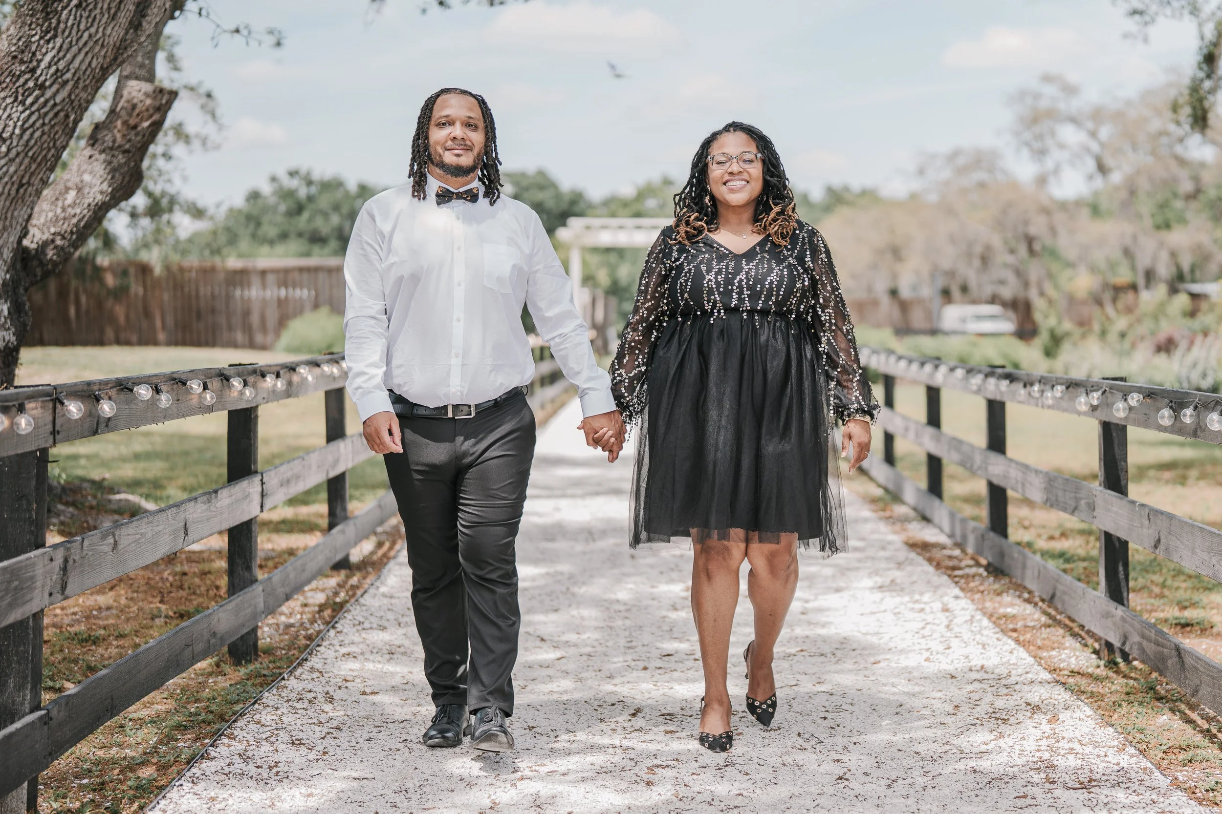 A man and woman holding hands and walking on a wooden pathway with string lights, trees, and a fence in the background, both smiling and dressed in semi-formal attire.