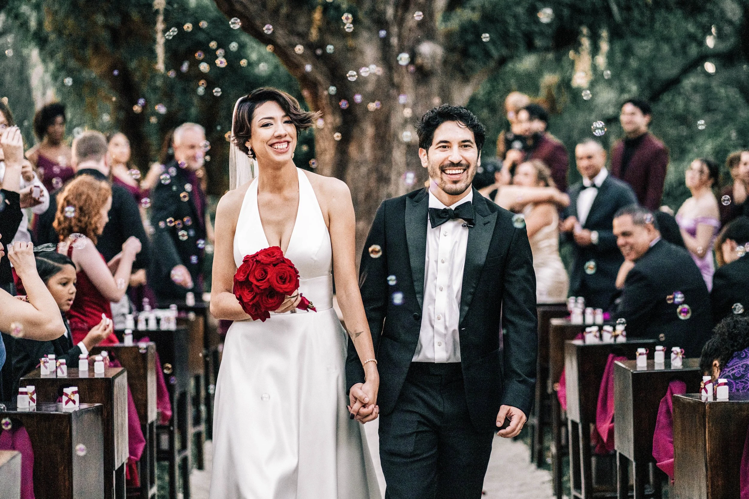 A wedding ceremony outdoors with a bride and groom holding hands and smiling, surrounded by guests and floating bubbles.