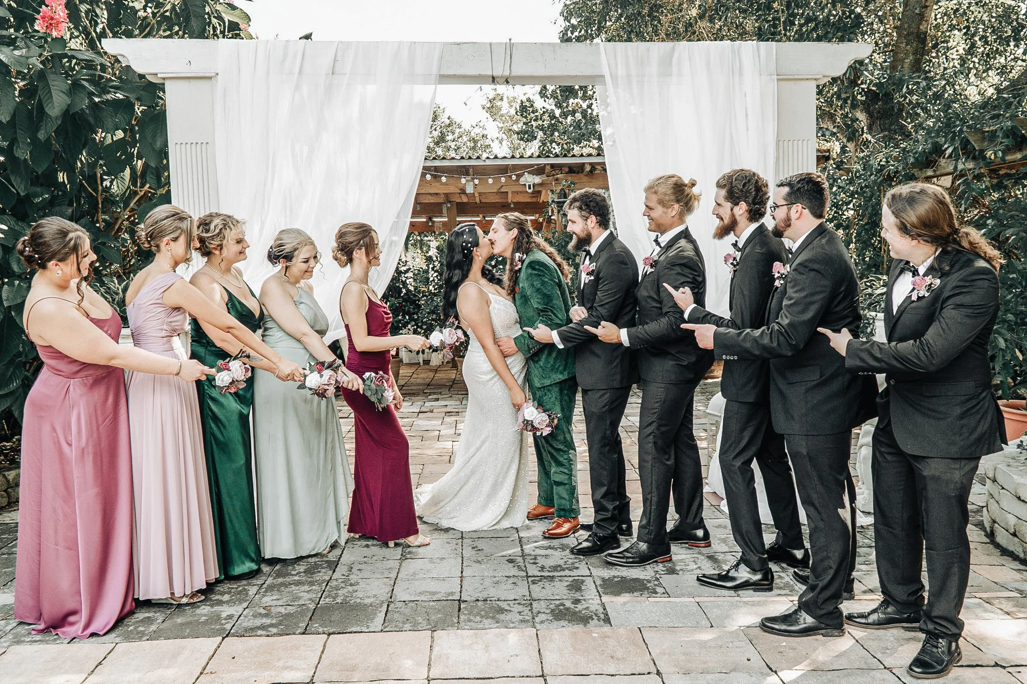 A wedding ceremony with the bride and groom kissing, surrounded by bridesmaids in colorful dresses and groomsmen in black suits, outdoors under a white canopy with trees in the background.