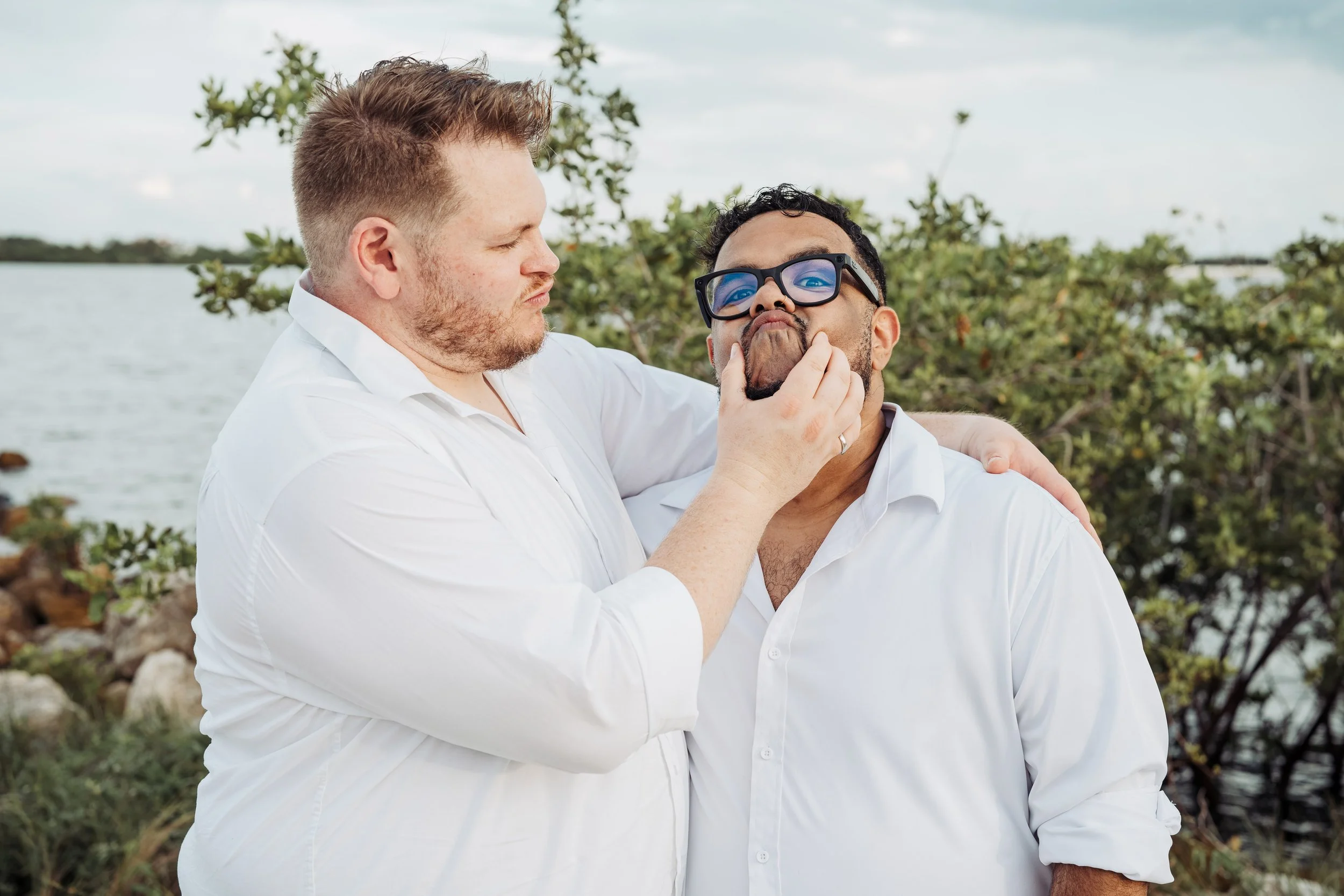 Two men outdoors near water, one with glasses and a beard, the other with short hair and no glasses, engaging in a playful moment where the man with glasses is being touched on the face by the other man.