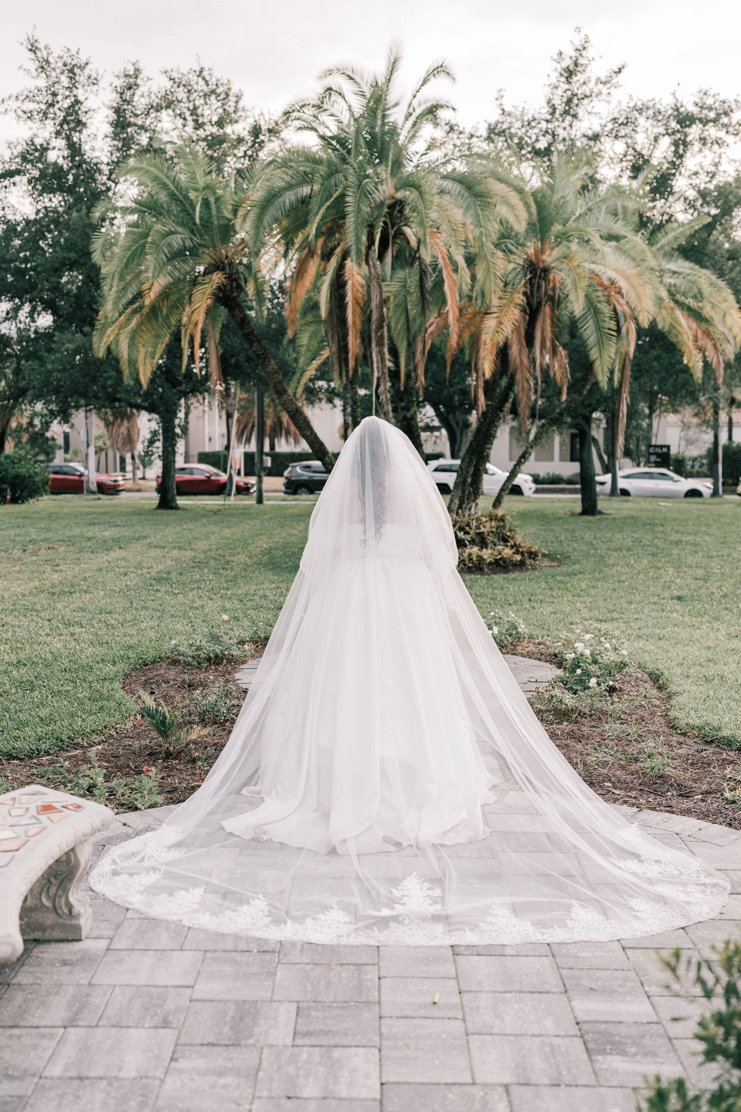A wedding dress with a long train on a paved garden path, with palm trees and parked cars in the background.