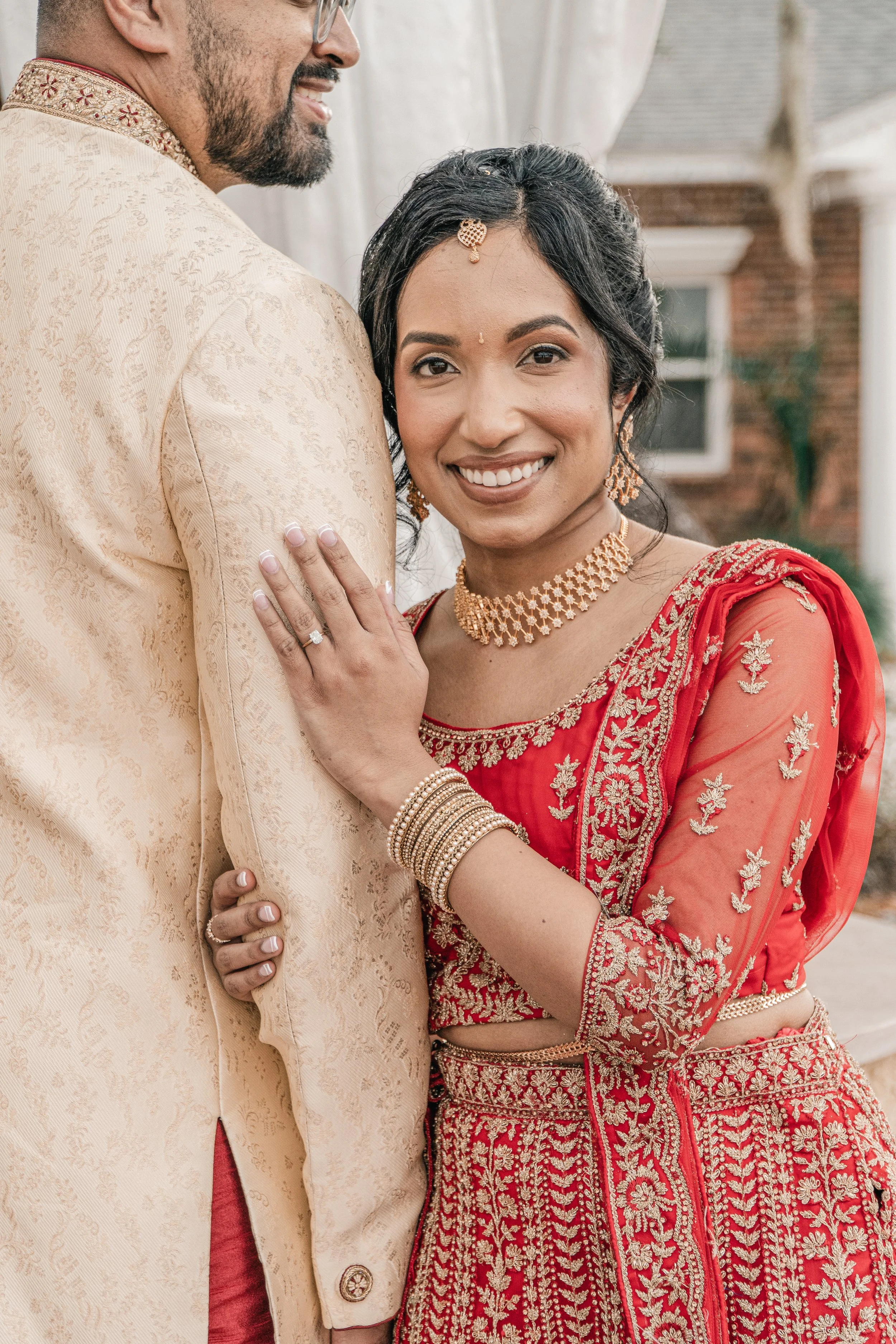 A happy couple dressed in traditional Indian wedding attire, with the woman wearing a red and gold embroidered saree and jewelry, and the man in an ivory sherwani with gold embroidery, outdoors with a house in the background.