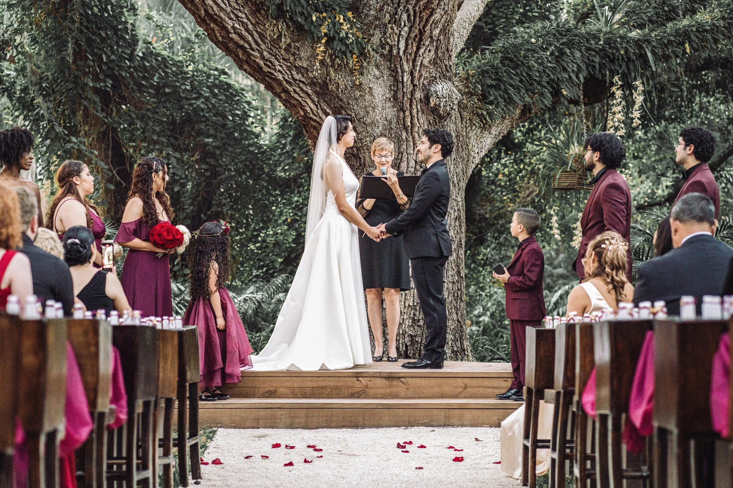 A wedding ceremony taking place outdoors under a large tree, with the bride and groom holding hands and exchanging vows in front of an officiant, surrounded by seated guests and bridesmaids and groomsmen.