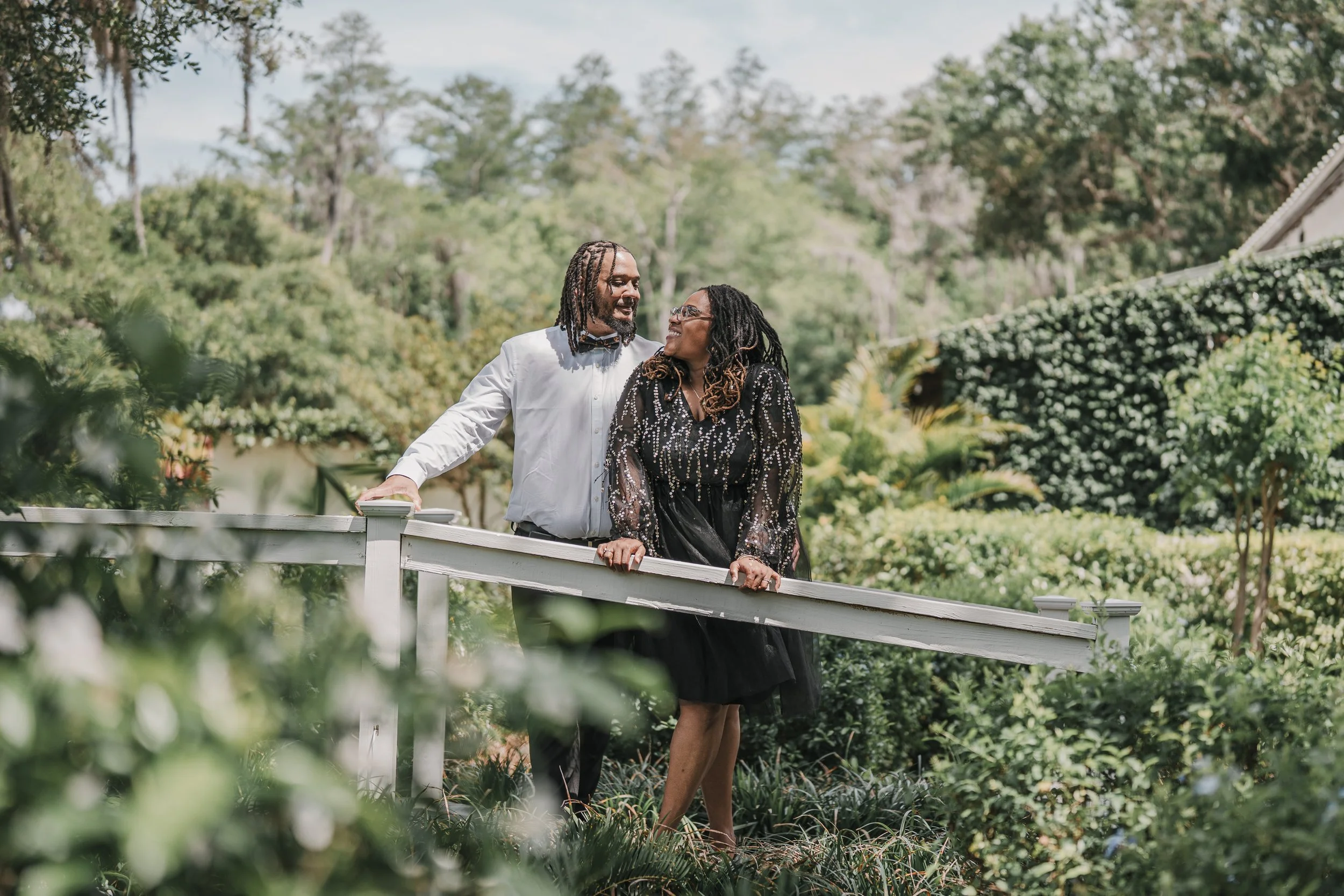 A couple standing outdoors on a white railing, smiling and looking at each other, surrounded by greenery and trees.