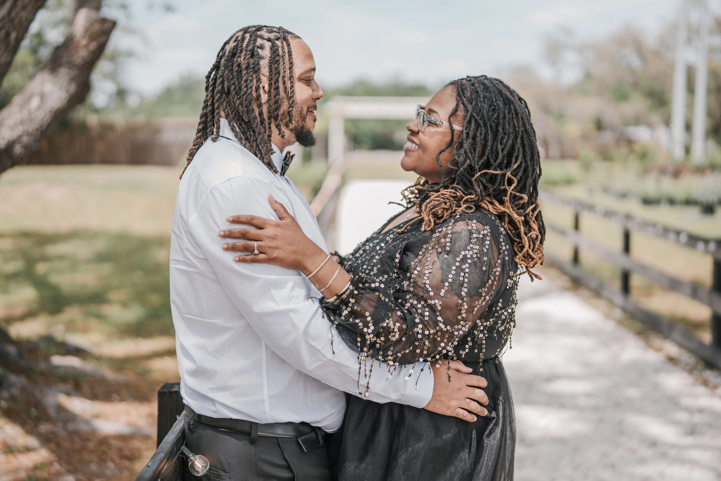 A couple standing close together outdoors, gazing into each other's eyes, smiling, with trees and a fence in the background.