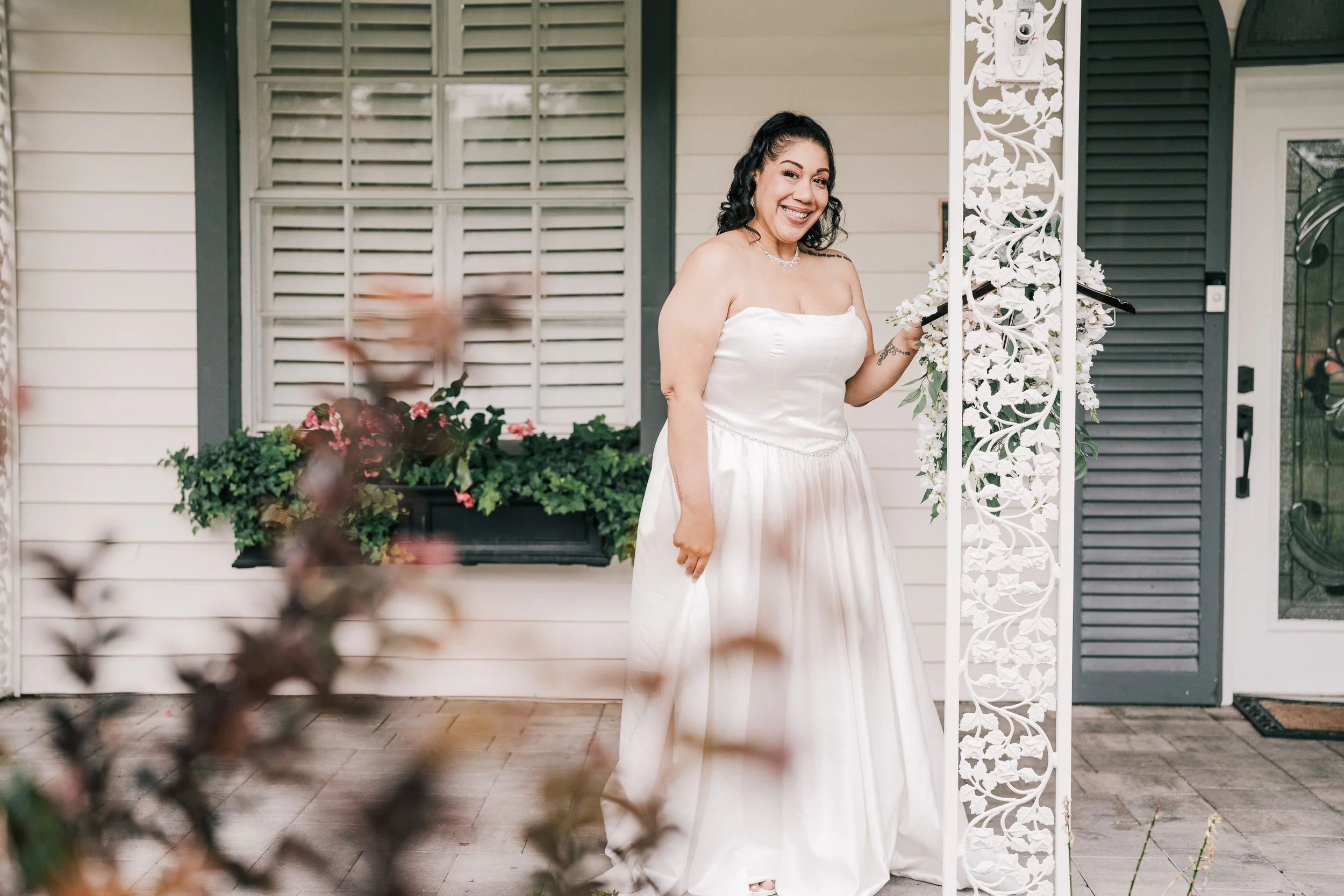 A woman in a white wedding gown smiling and holding a black clutch, standing next to a decorative white floral pillar outside a house with beige siding, black shutters, and potted pink flowers.