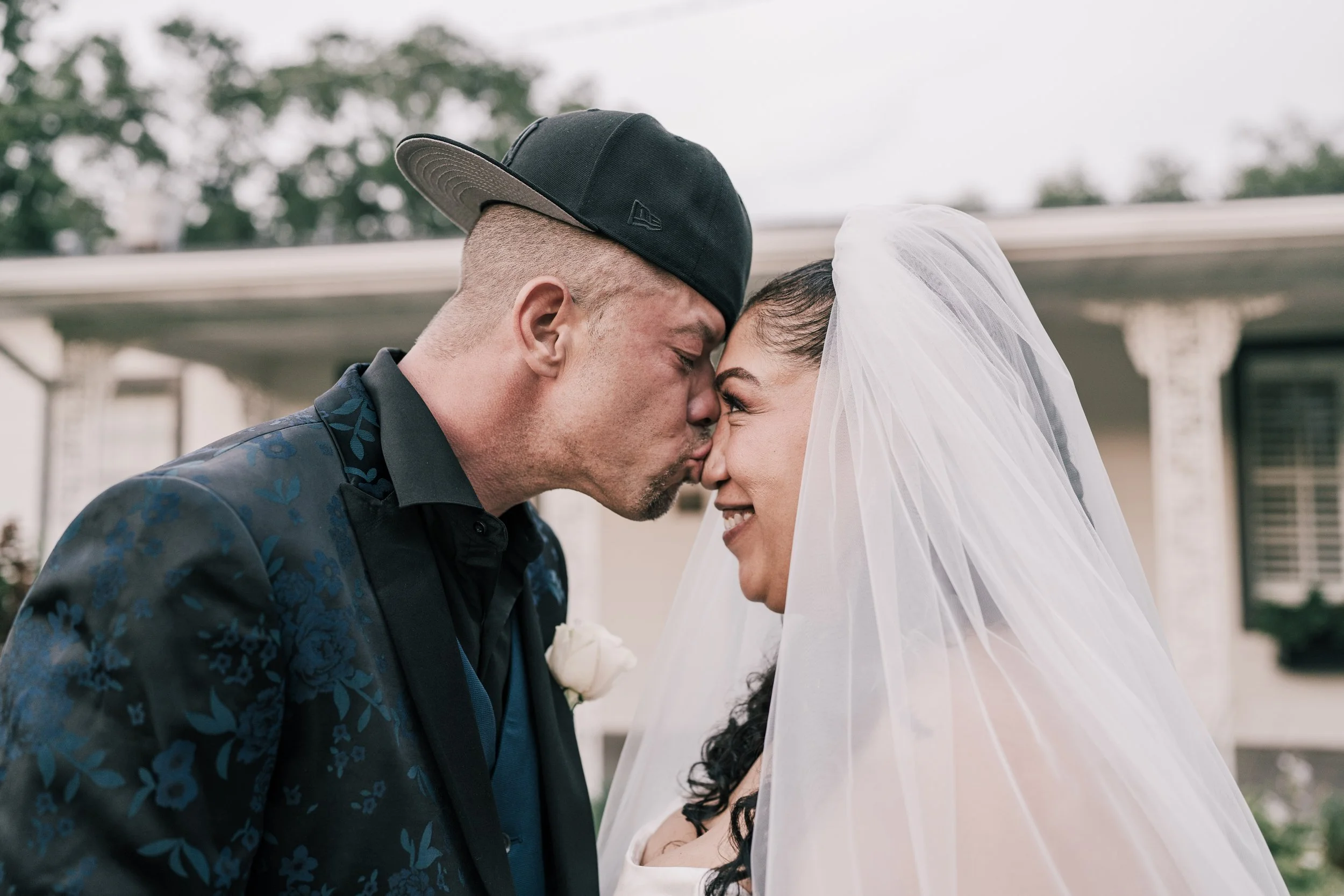 A bride and groom share a kiss during their wedding ceremony outdoors, with green trees in the background.