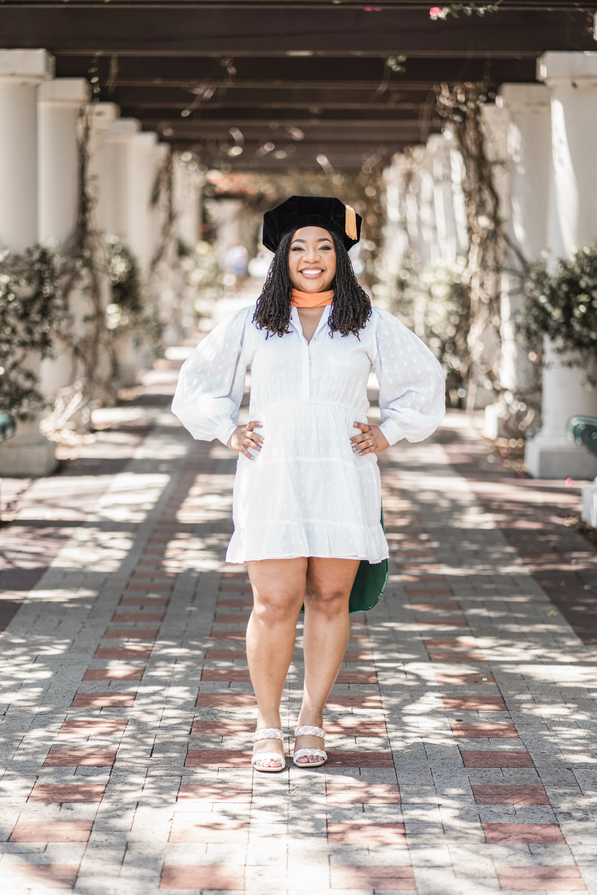 A young woman dressed in a white dress standing outdoors on a brick pathway, smiling. She is wearing a black academic cap and holding her hands on her hips. The background features white columns and greenery.