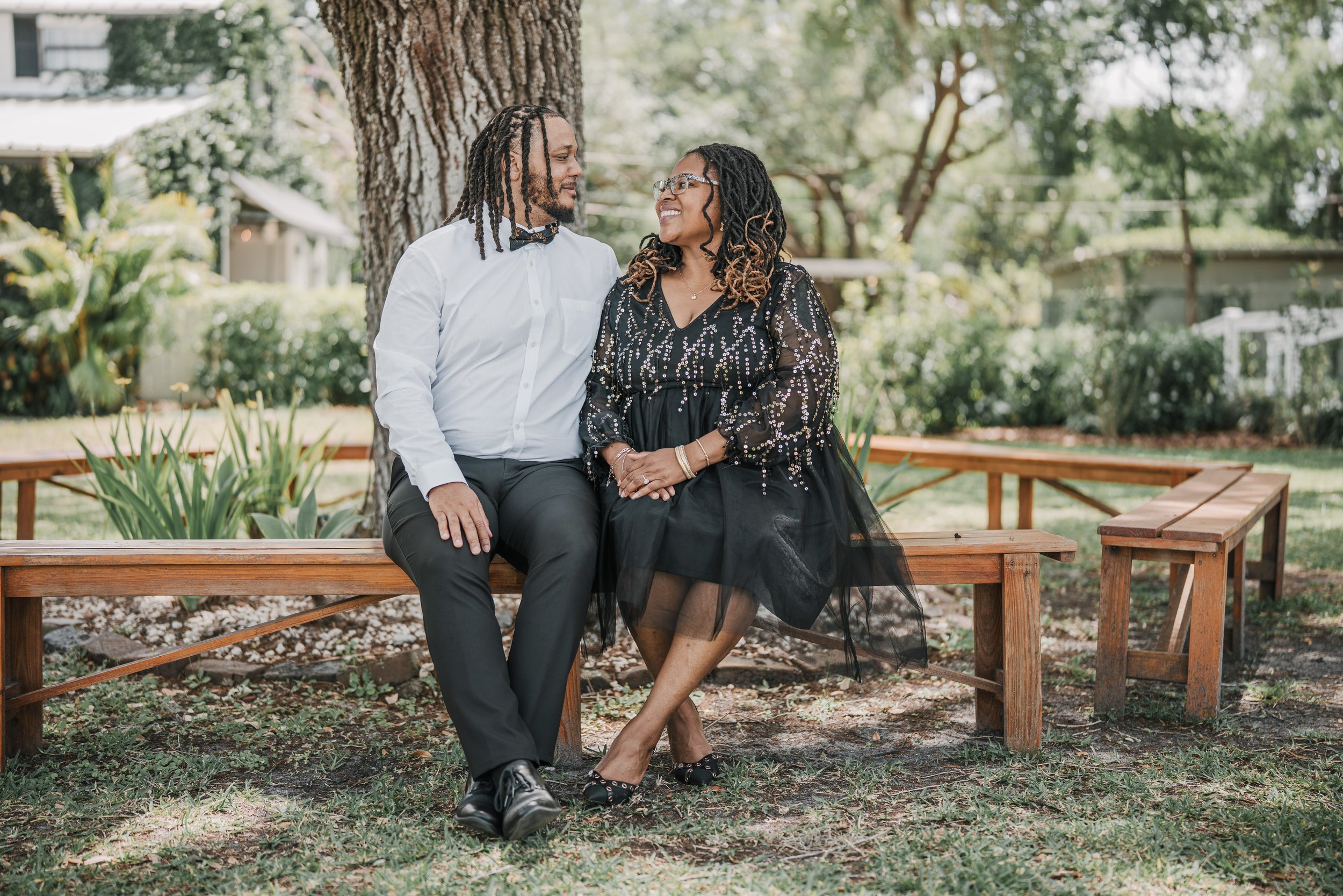 A man and woman sitting on a wooden bench under a large tree in a yard, smiling and looking at each other, with greenery and a house in the background.