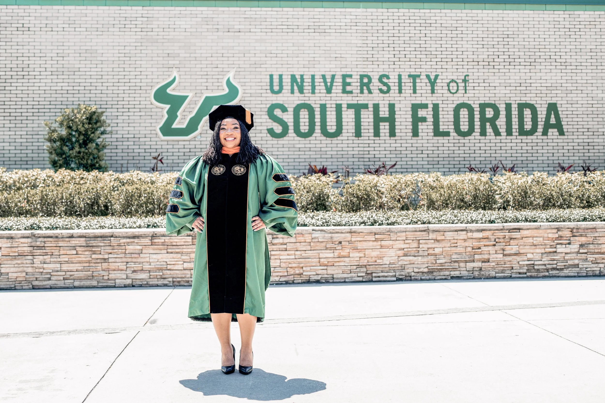 A woman dressed in graduation regalia, standing in front of a sign that reads 'University of South Florida' on a brick wall. She is smiling and posing for a photo.